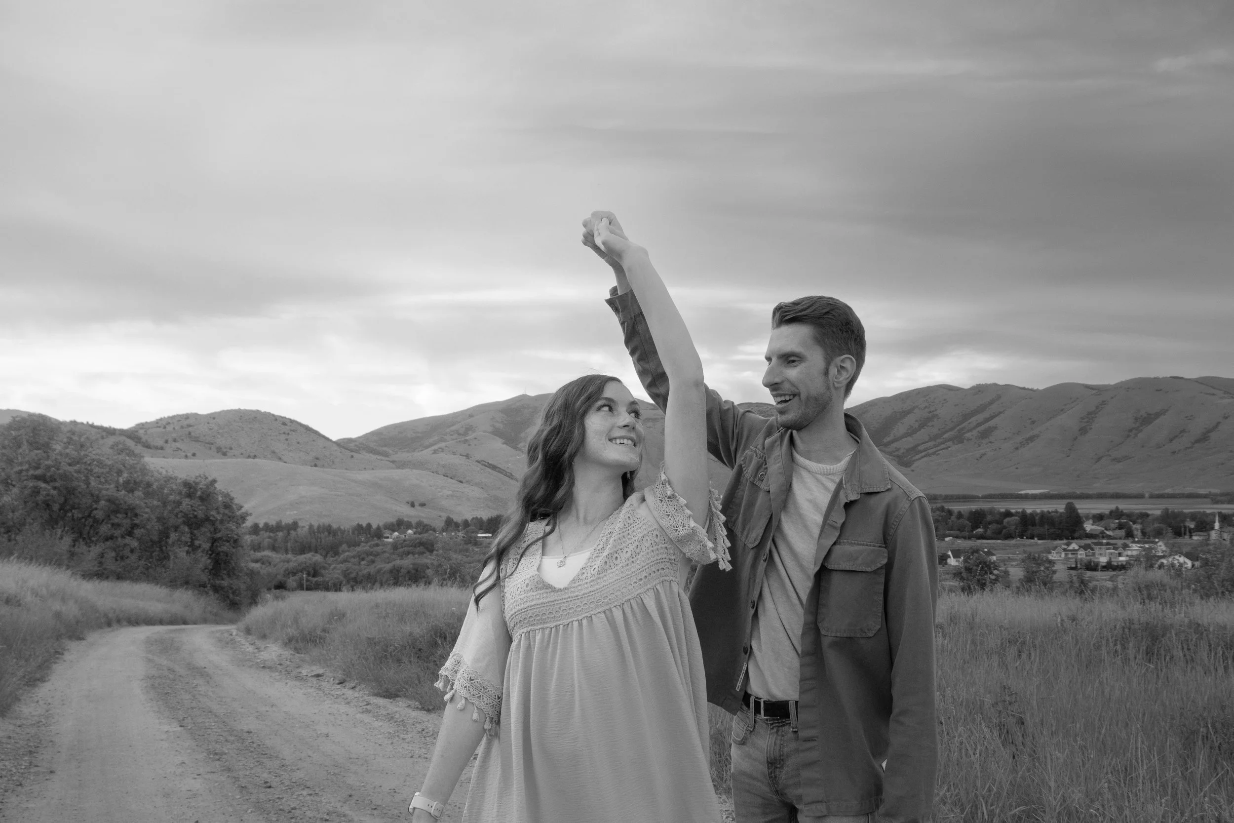 A black and white photo of a smiling couple holding hands high in the air outdoors with rolling hills and a cloudy sky in the background.