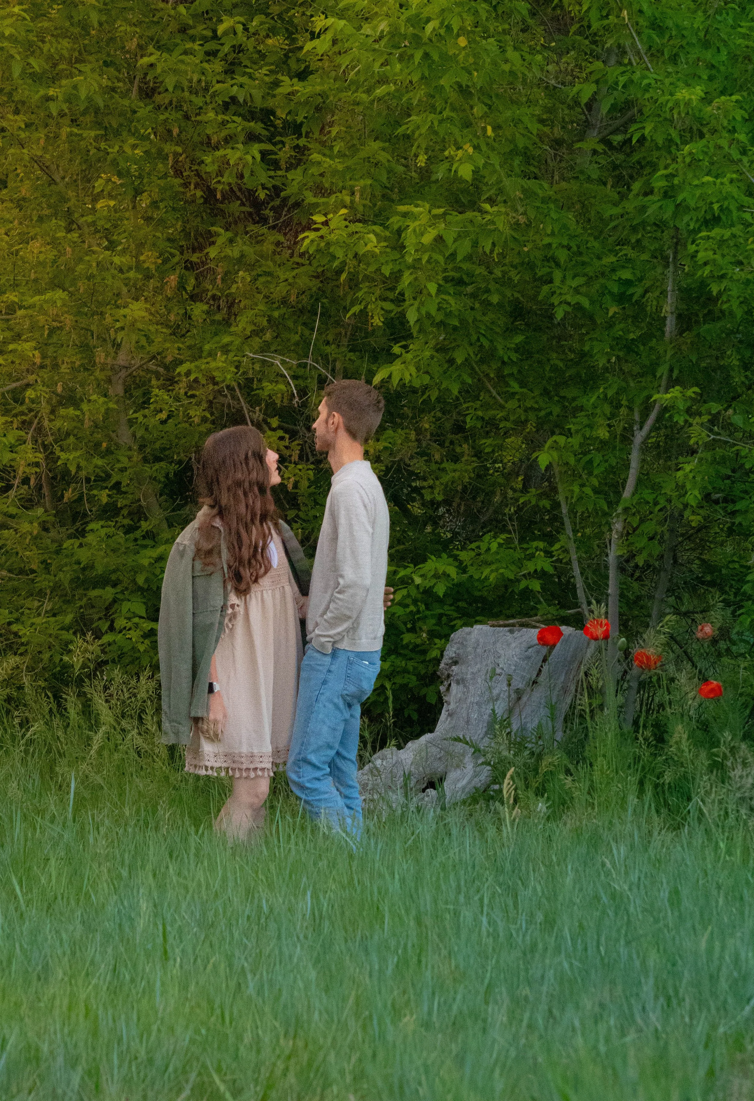 A man and woman standing closely together outdoors, looking into each other's eyes, with lush green trees and red flowers in the background.