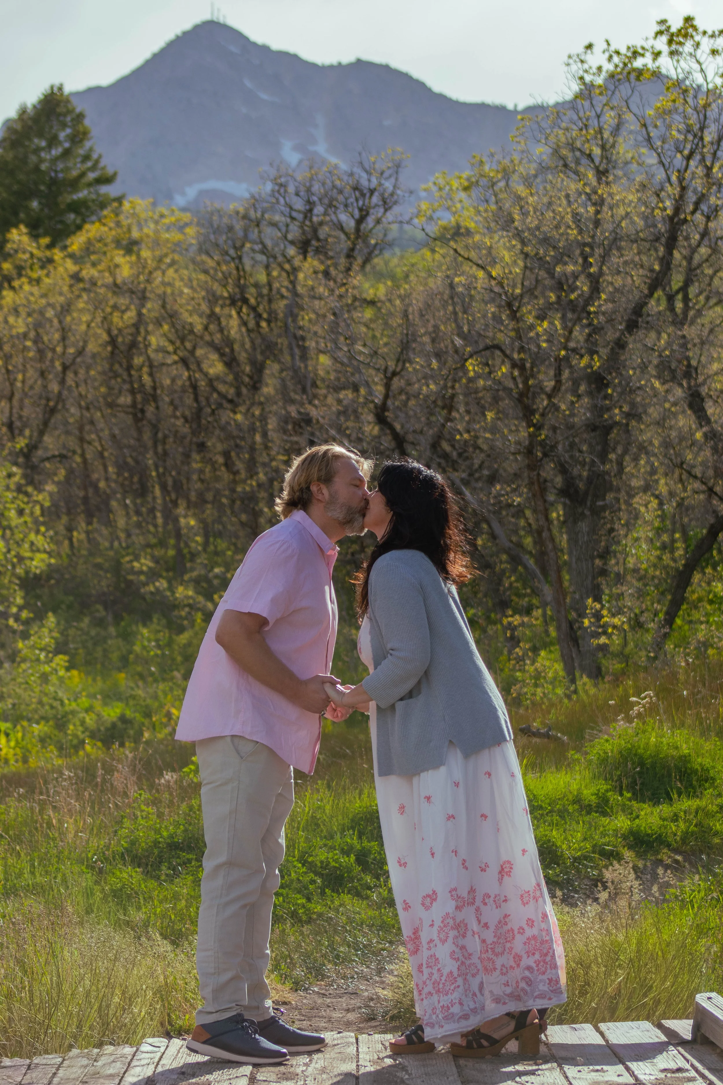 A couple is holding hands and kissing outdoors on a wooden bridge, with trees and mountains in the background on a sunny day.