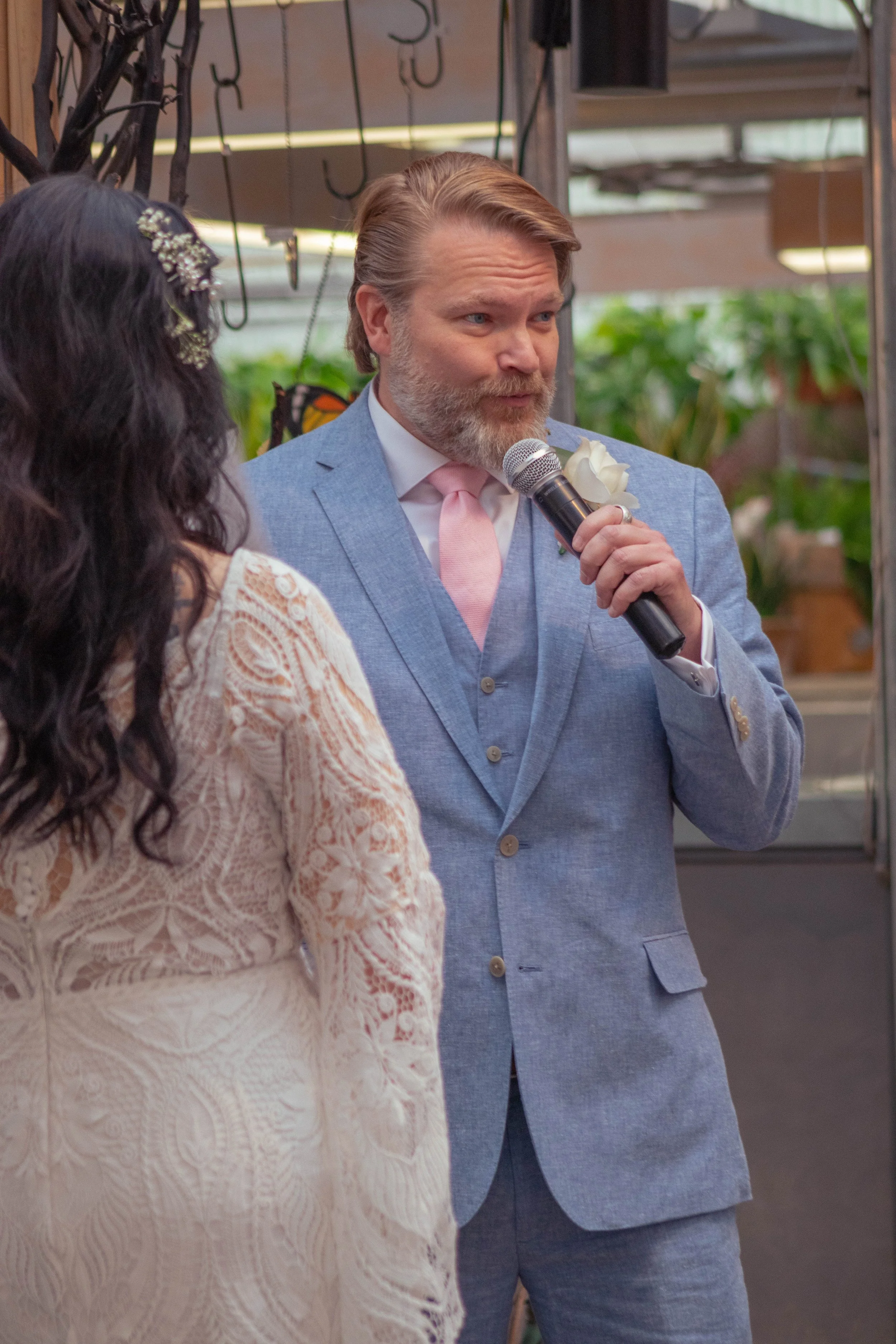 A man in a light blue suit with a pink tie and white flower boutonniere speaking into a microphone at a formal event, standing next to a woman in a cream lace dress with dark curly hair and floral hair accessory, indoors with plants and hooks hanging