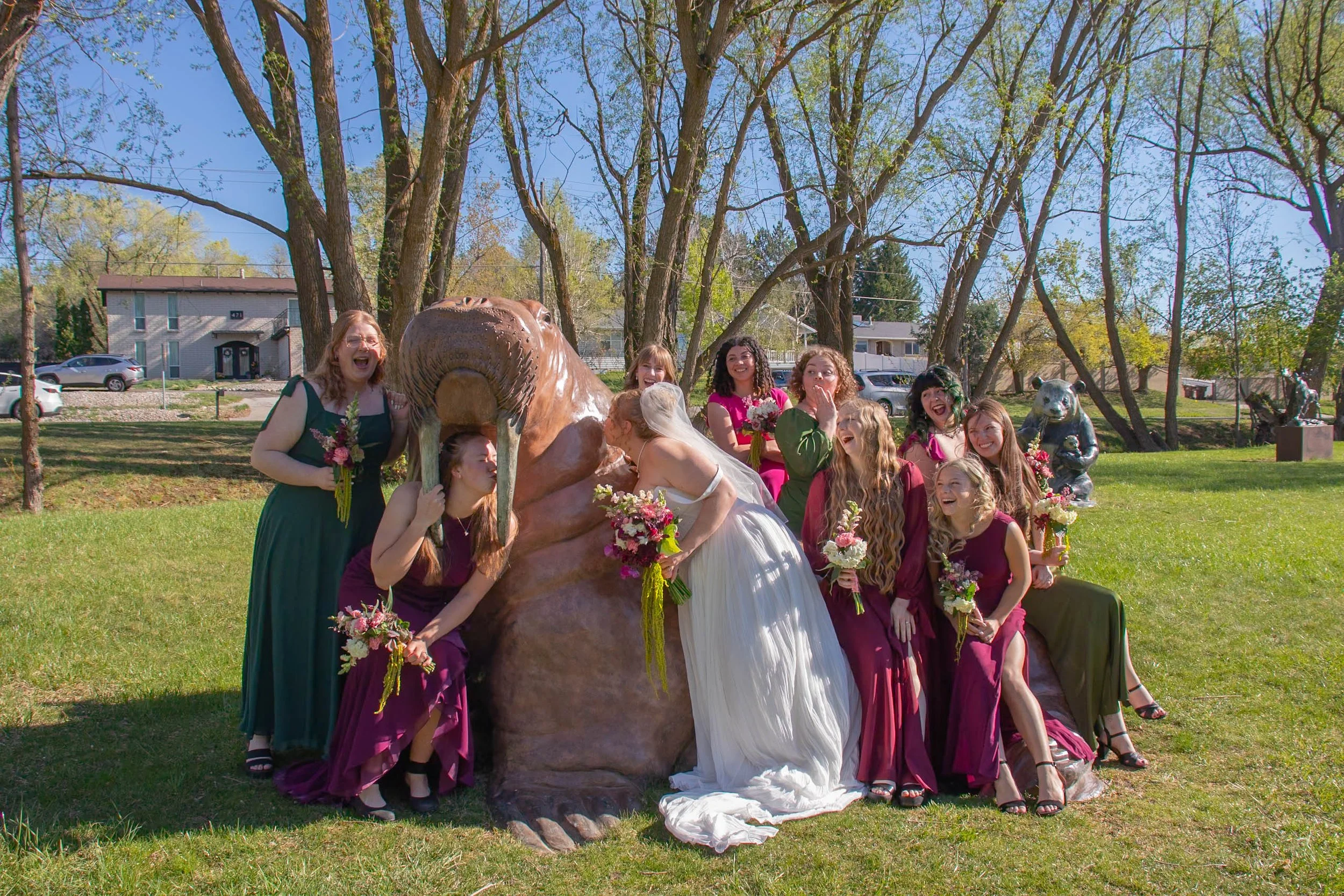 A group of women and children dressed in colorful dresses, gathered around and kissing a bronze statue of a large bird, possibly a heron or stork, in a park with trees and houses in the background.