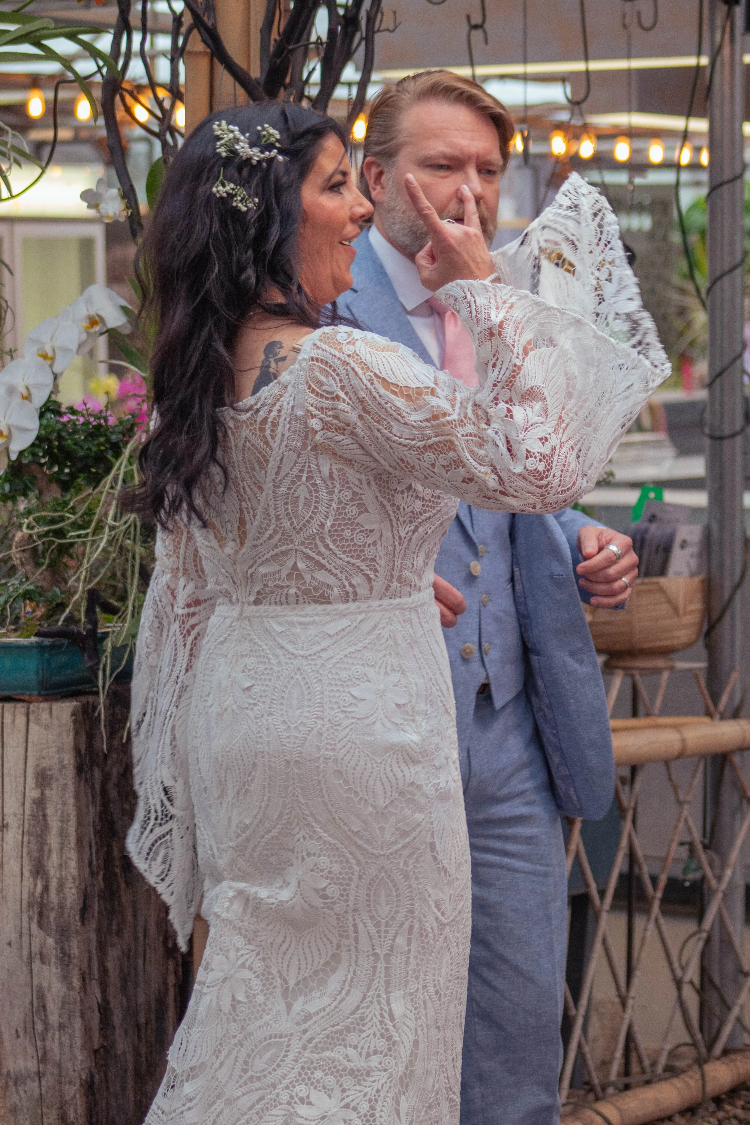 A woman in a white lace dress with flowers in her hair is touching the nose of a man in a blue suit with a pink tie, indoors with decorative plants and lights.