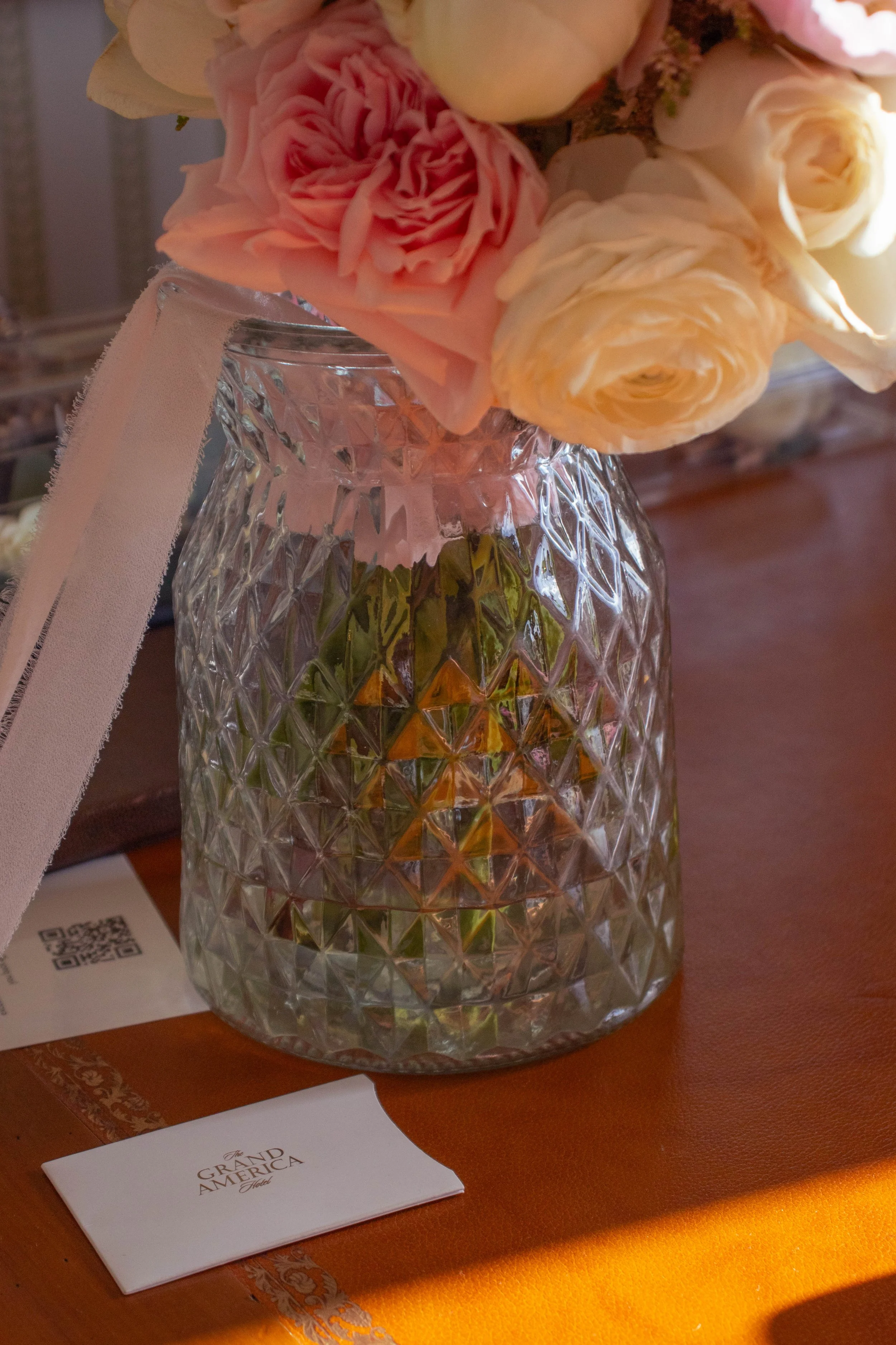 A glass vase with a diamond pattern holds a bouquet of pink and peach roses. The vase is on a table beside a white card with black text that reads 'The Grand America Hotel' and some other items.