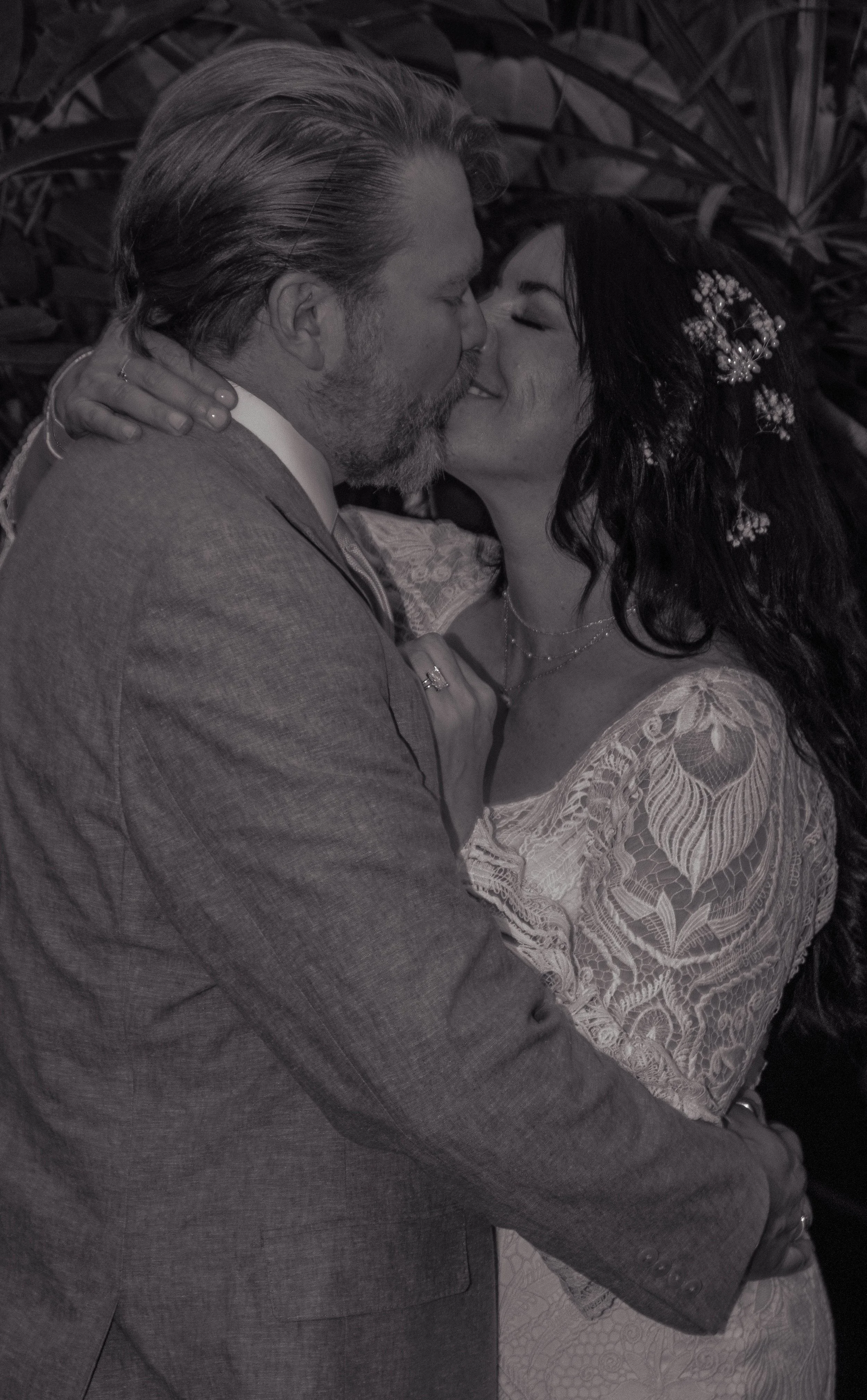A couple sharing a kiss, with the woman wearing a lace dress and floral hair accessory, and the man in a suit, in an outdoor setting with foliage in the background.