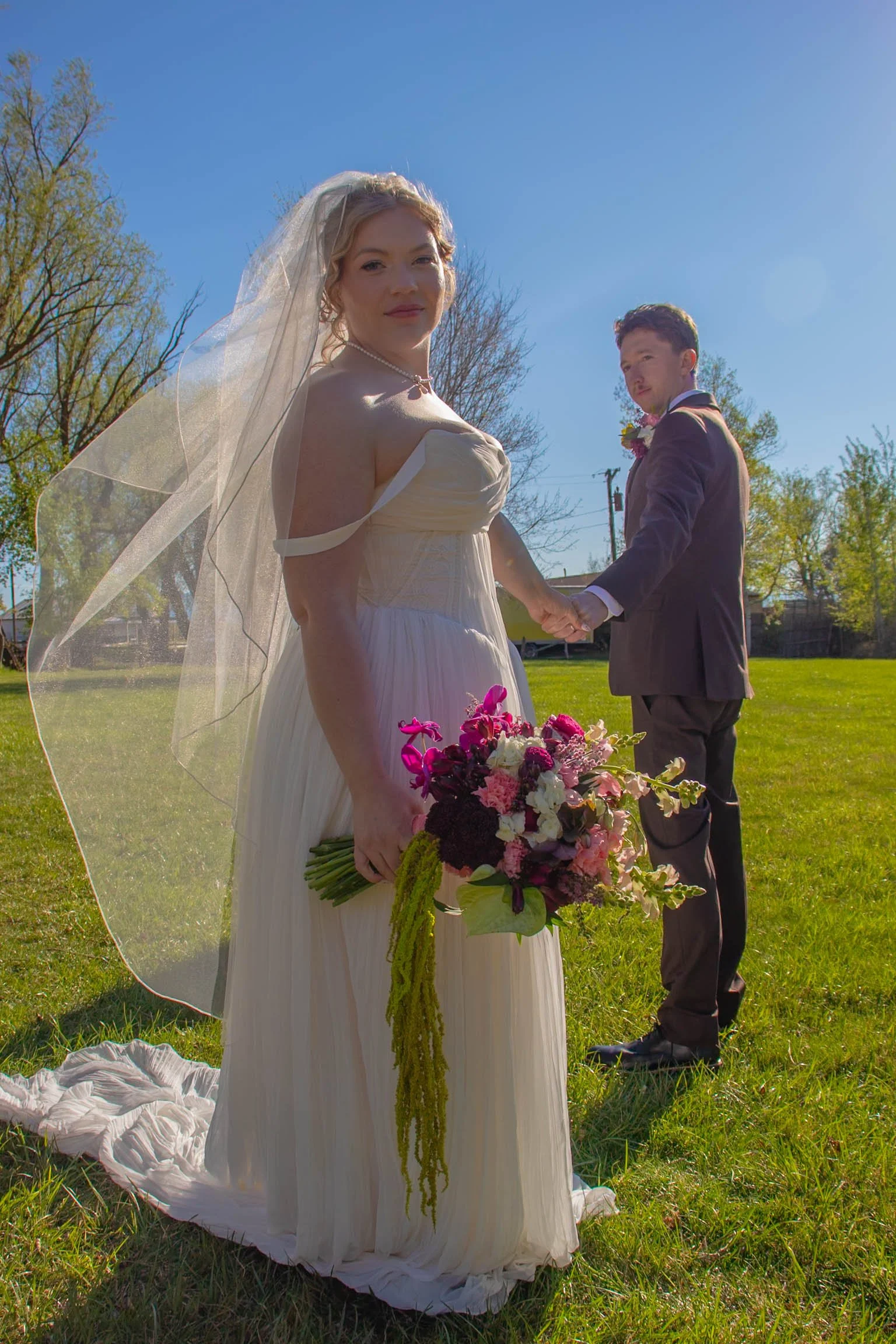 A bride in a white strapless wedding gown holding a bouquet of pink, purple, and white flowers, standing outdoors on a sunny day with a man in a suit in the background.