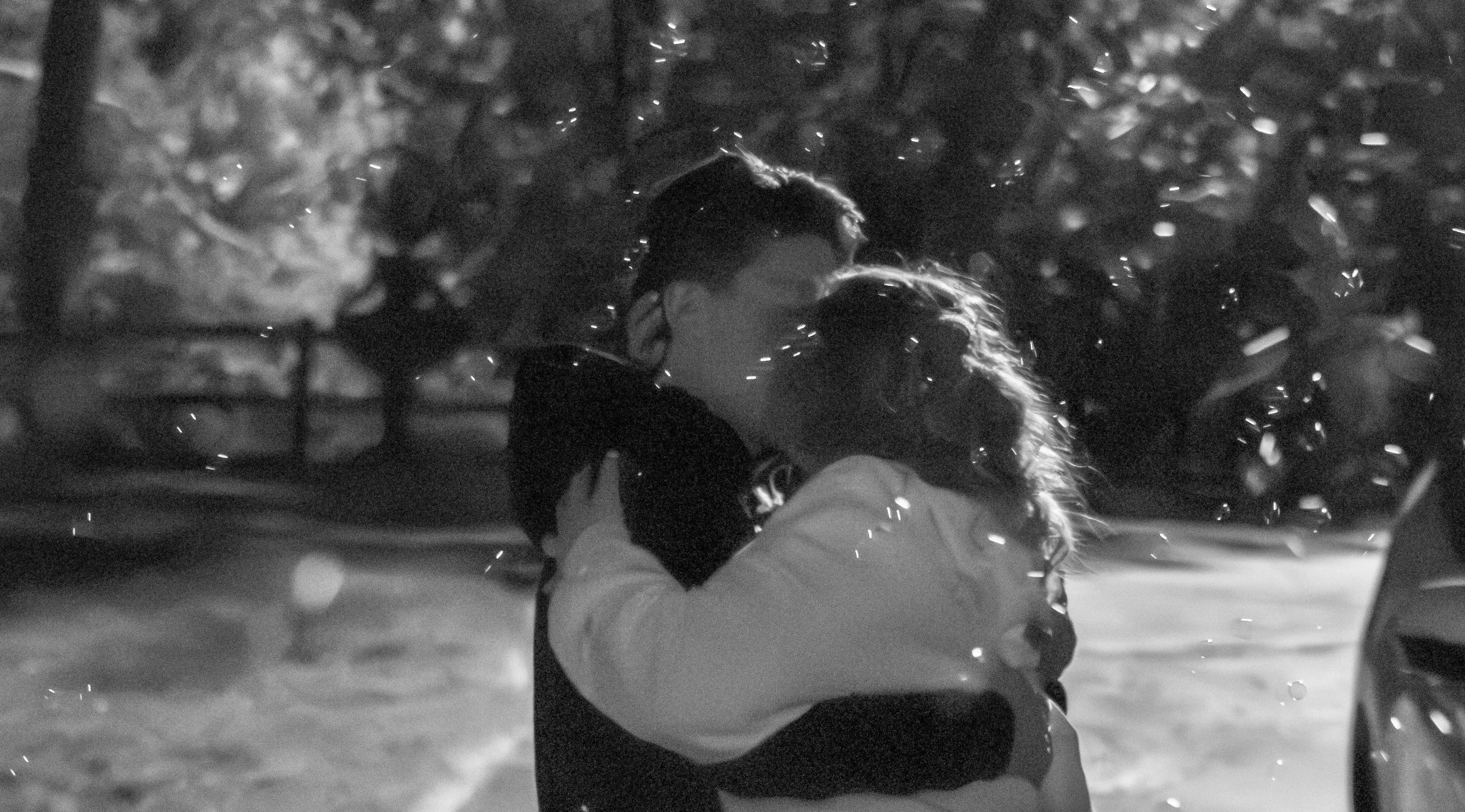 A person with curly hair is embracing another person in a rainy outdoor setting at night, with raindrops visible in the air.