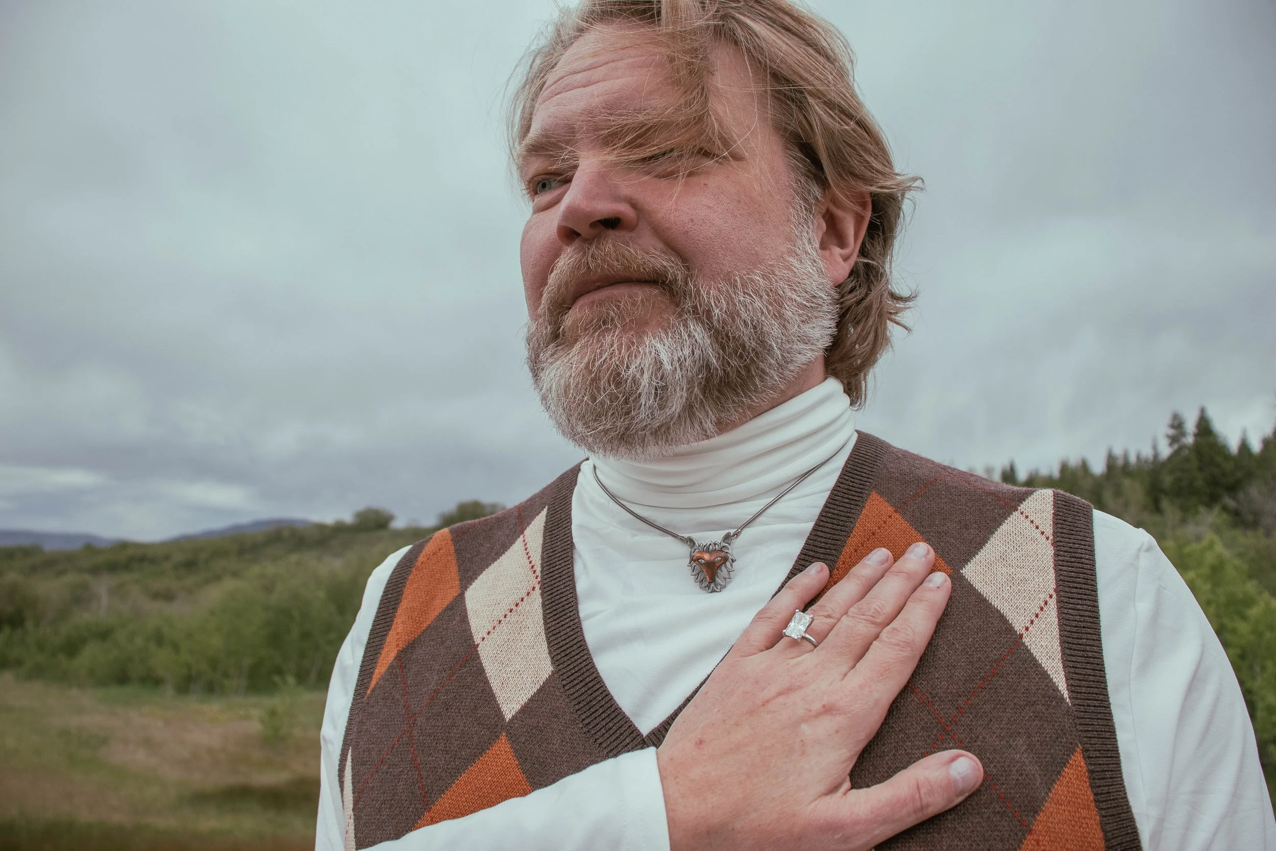 A middle-aged man with a beard and long, light-colored hair standing outdoors under an overcast sky, wearing a white turtleneck shirt and a brown argyle sweater vest with orange and beige diamonds, with his right hand over his heart and wearing a lio
