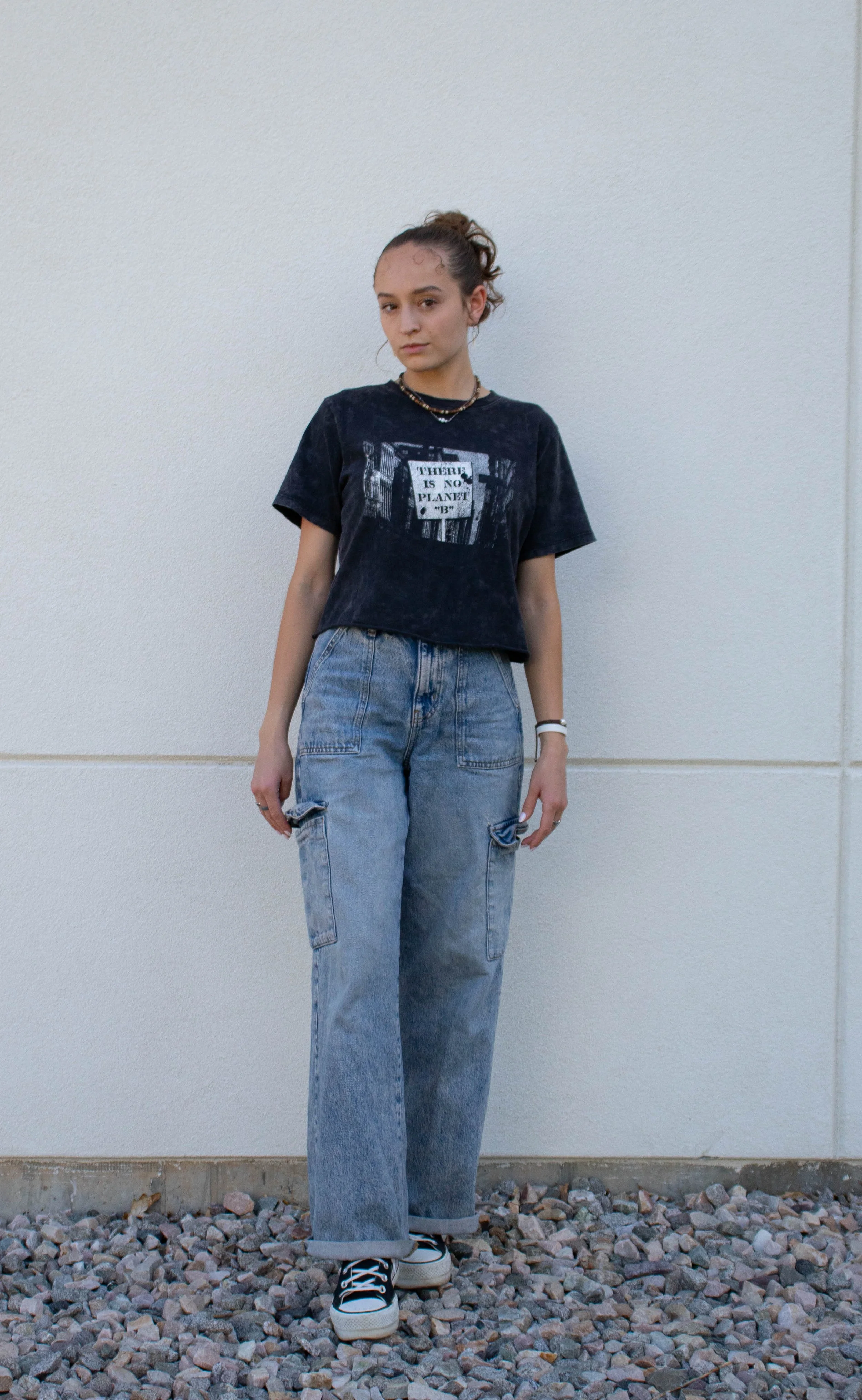 Young woman standing on rocks against a plain wall, wearing a black graphic T-shirt, baggy jeans, and sneakers.