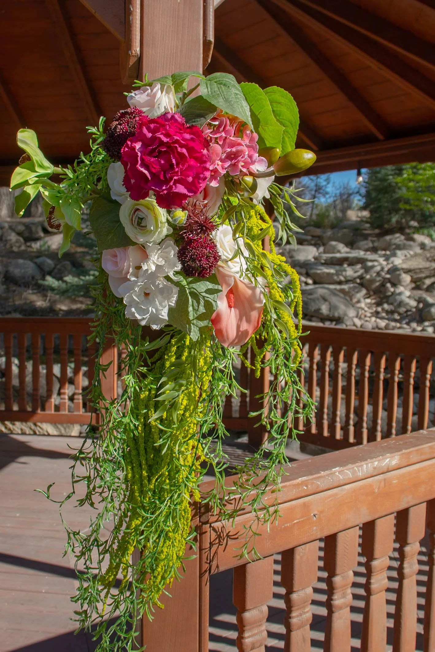 A hanging floral arrangement with pink, white, and green flowers and greenery, attached to a wooden deck railing.