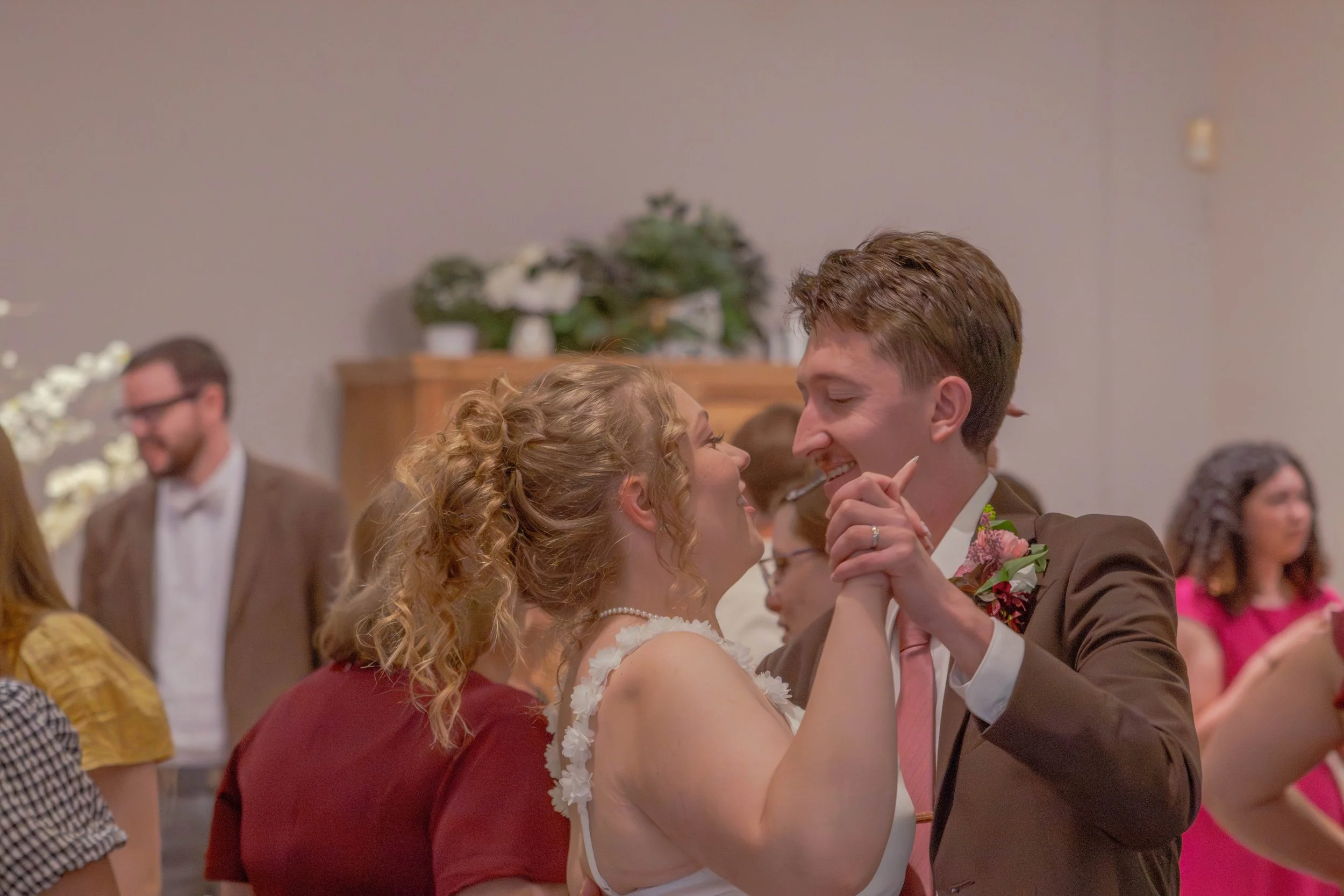 A couple is dancing closely at a wedding, smiling and gazing into each other's eyes, surrounded by other guests in the background.