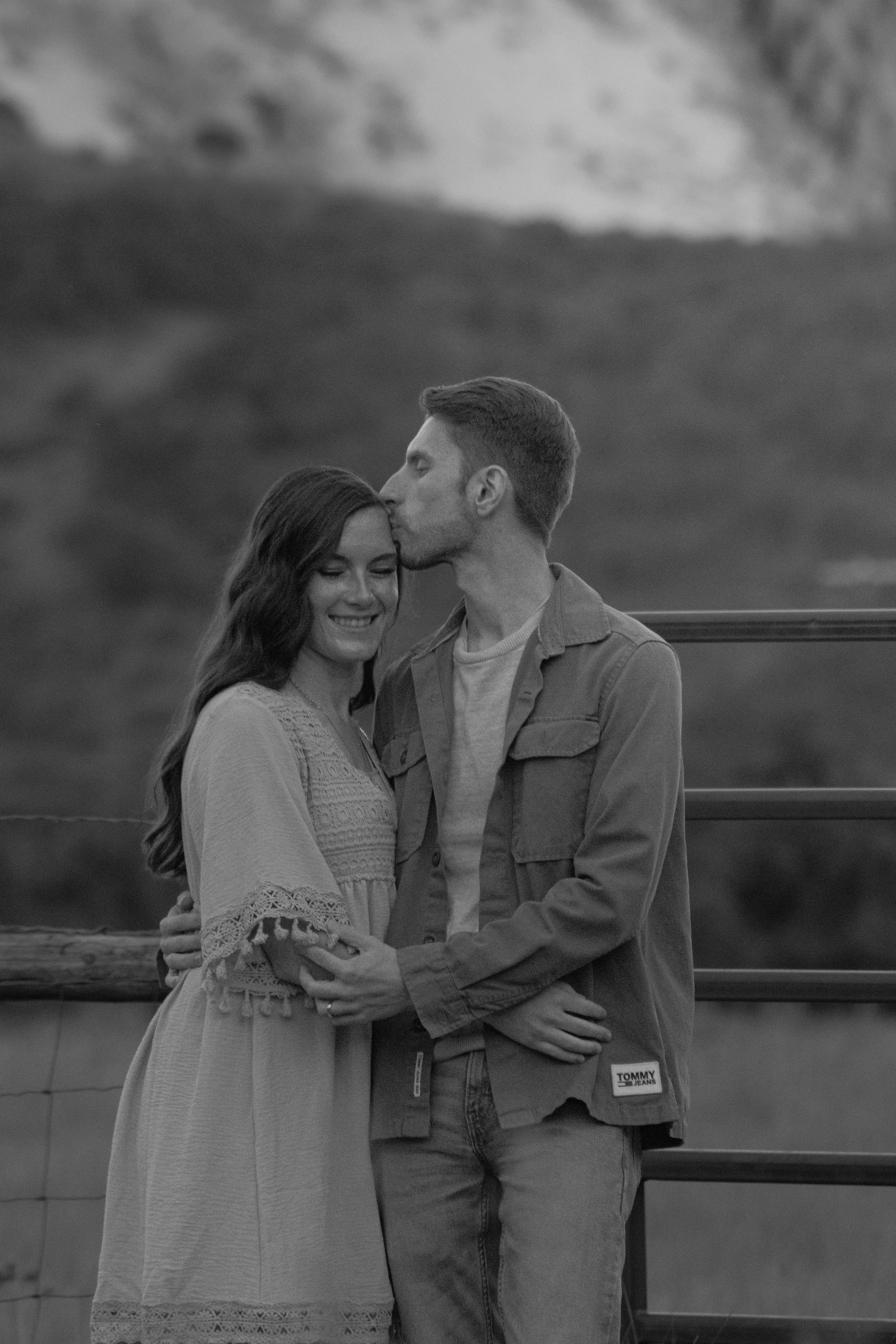 A black and white photo of a couple standing outdoors, with the man gently kissing the woman's forehead. They are embracing each other with the woman's eyes closed and smiling, and the man looking down at her. In the background, there is a cloudy sky