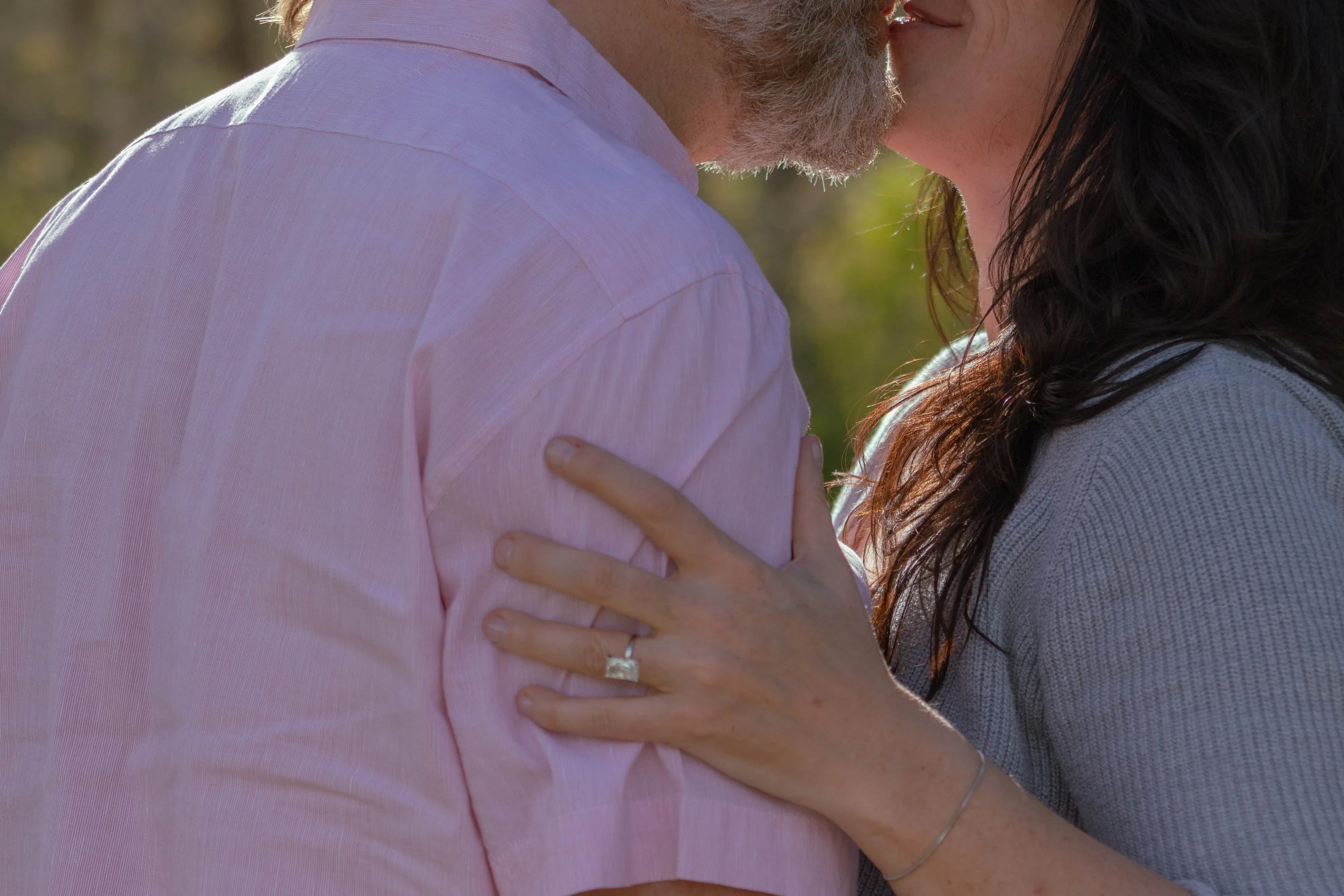 A close-up of a couple embracing and kissing outdoors, with sunlight highlighting their faces and arms, and a blurred natural background.