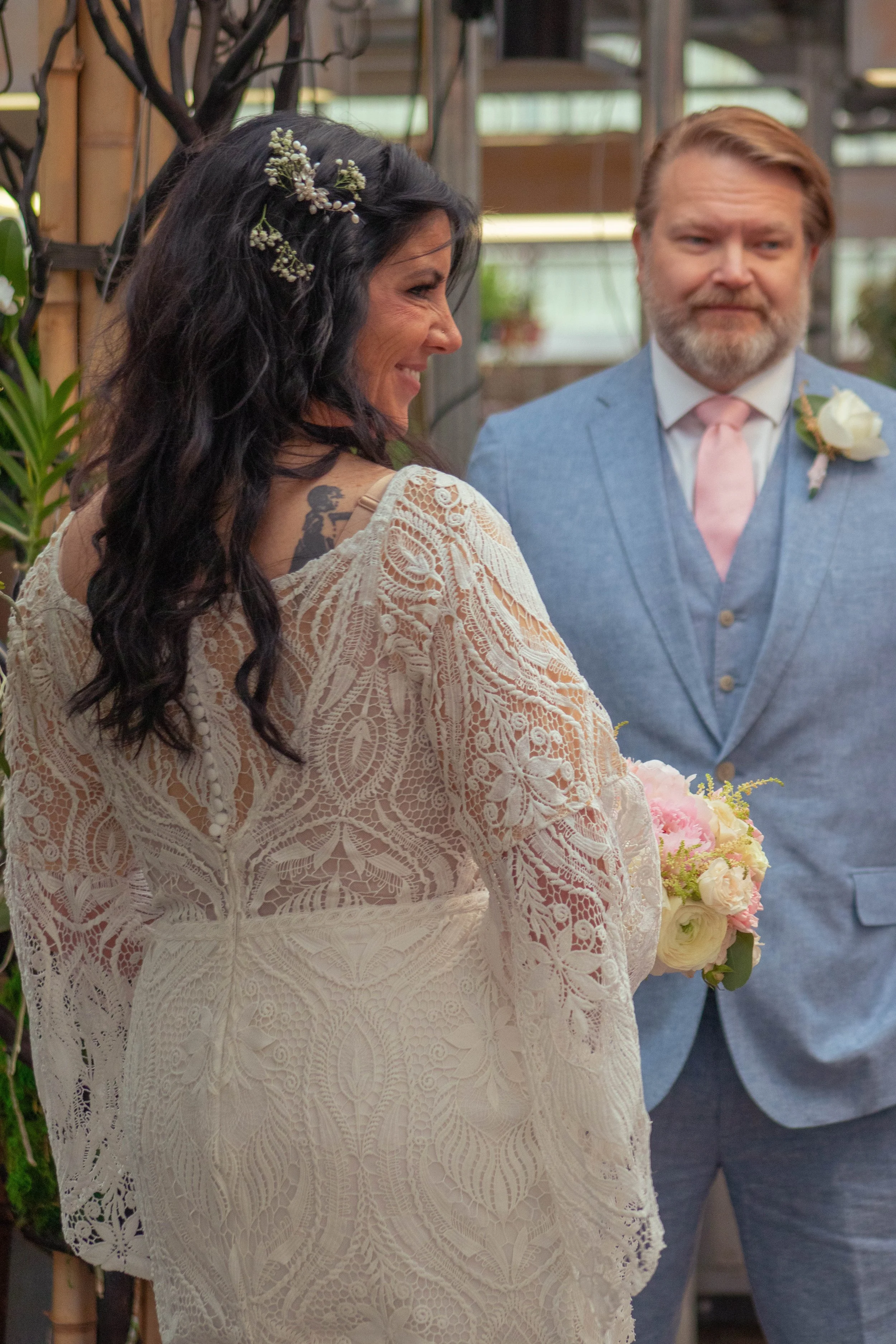 A bride with dark hair styled with small white flowers and a groom with a beard and hairstyle in a blue suit with a pink tie and boutonniere, standing at a wedding ceremony in a greenhouse setting.