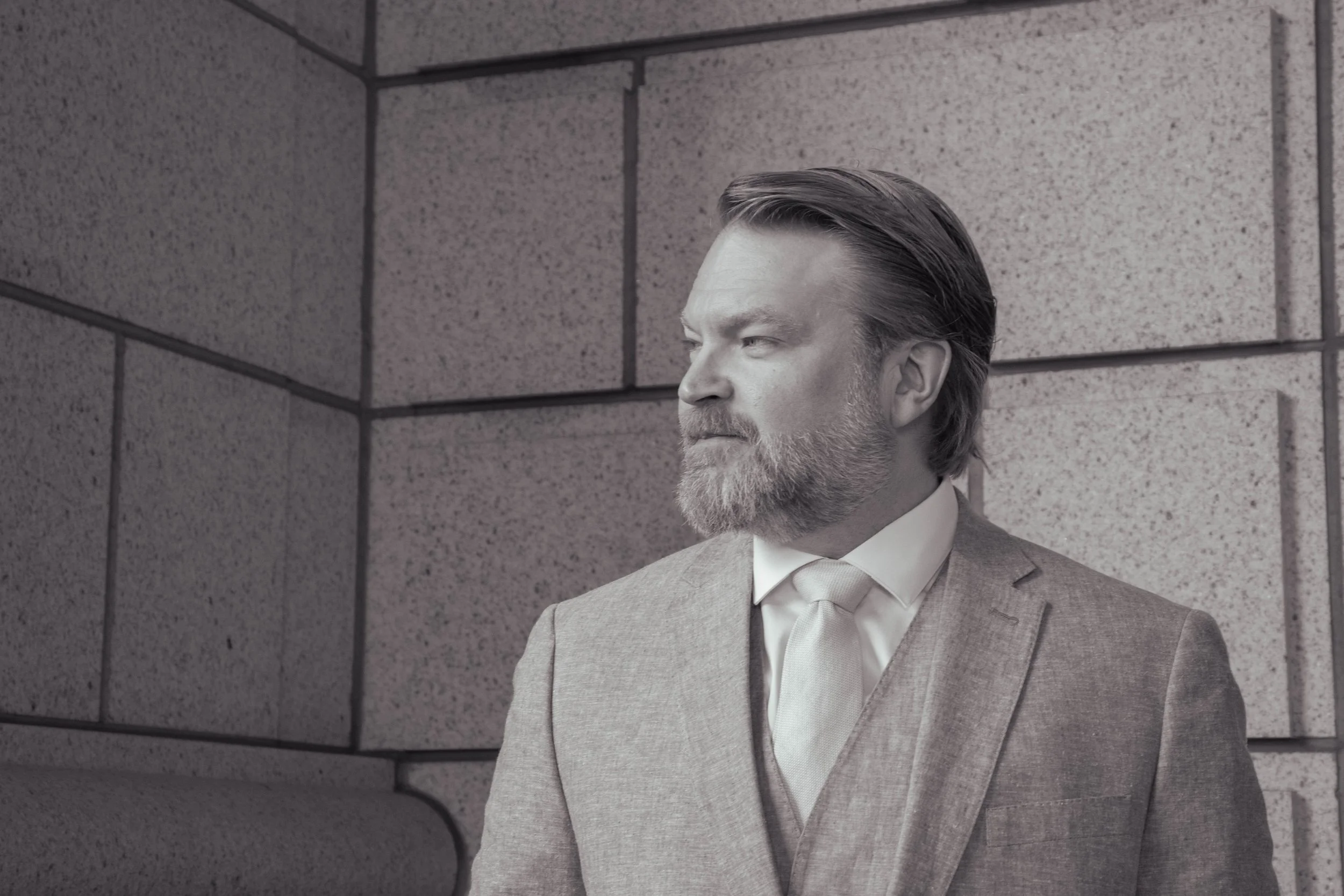 A man with gray hair and beard in a gray suit stands against a gray brick wall, looking to his right.