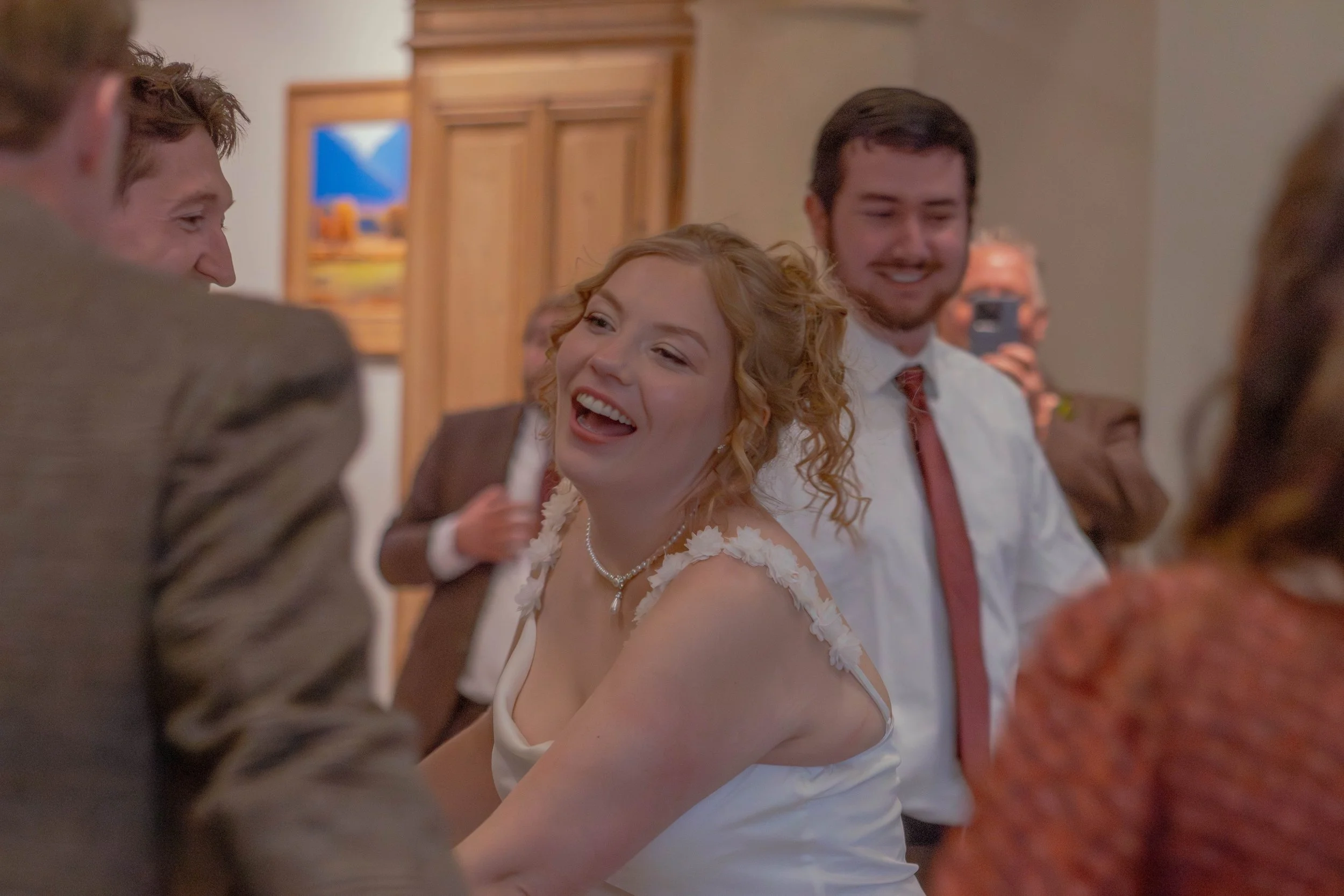 A group of people, including a woman with curly hair and a white dress, are dancing and laughing at a celebration in an indoor setting.