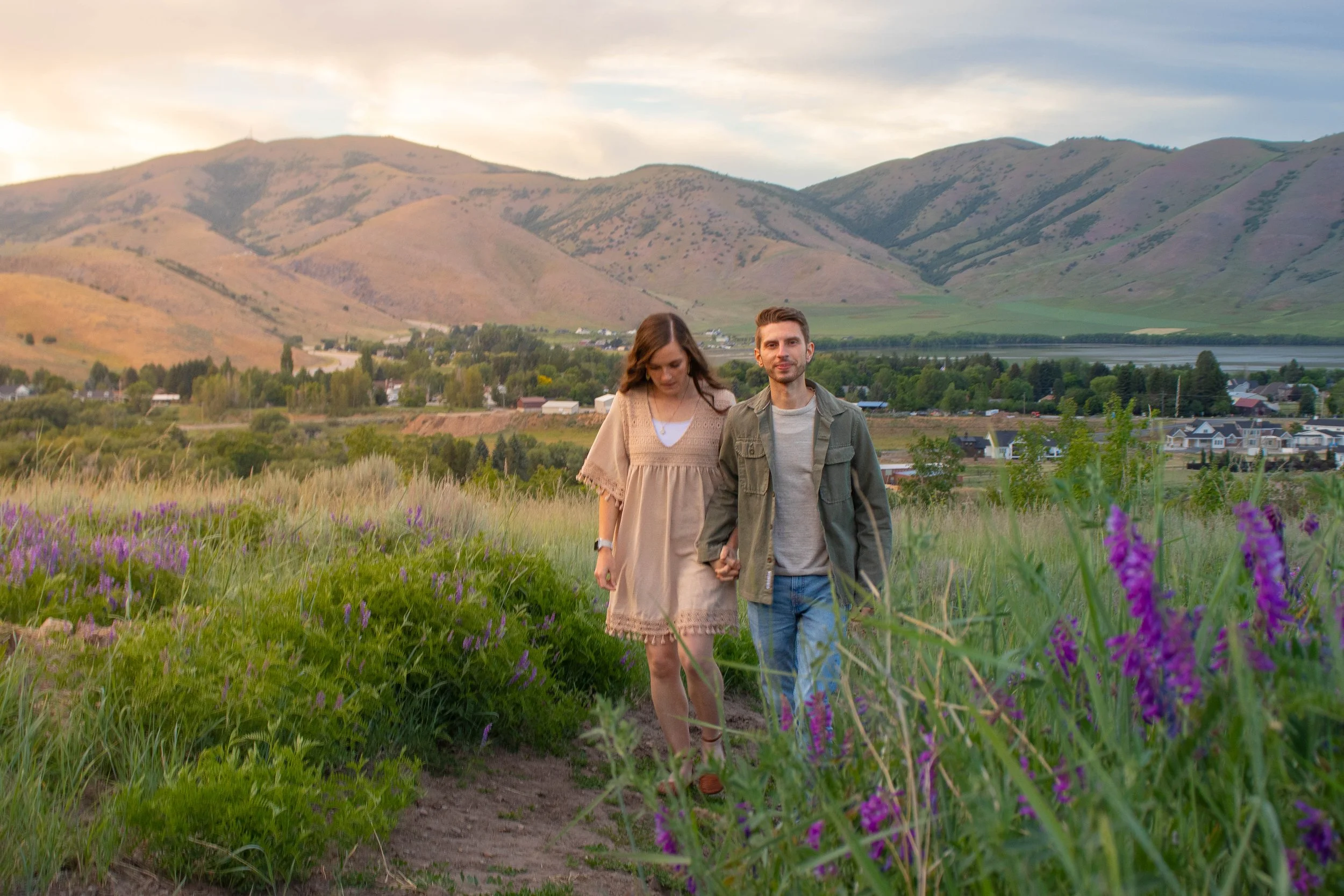 A young couple holding hands walking on a dirt trail through purple wildflowers with mountains in the background during sunset.