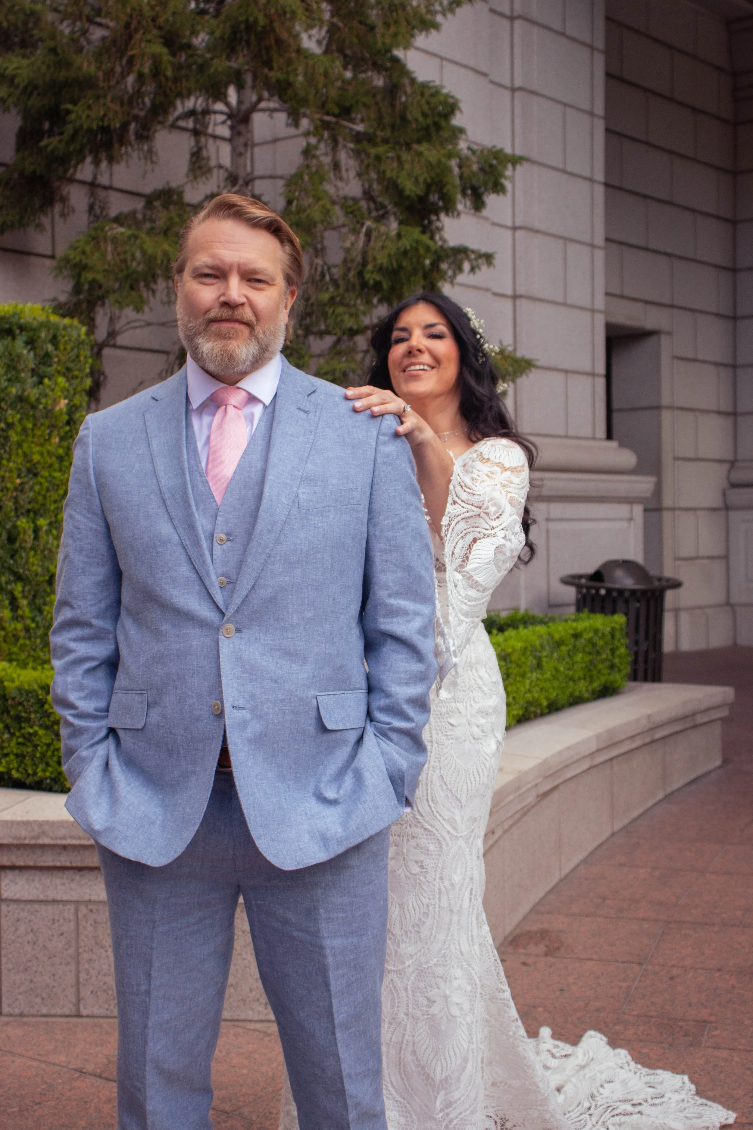 A man in a light gray suit with a pink tie stands in front of a woman in a white lace wedding dress. The woman is smiling and resting her hand on the man's shoulder. There are trees and a stone building in the background.
