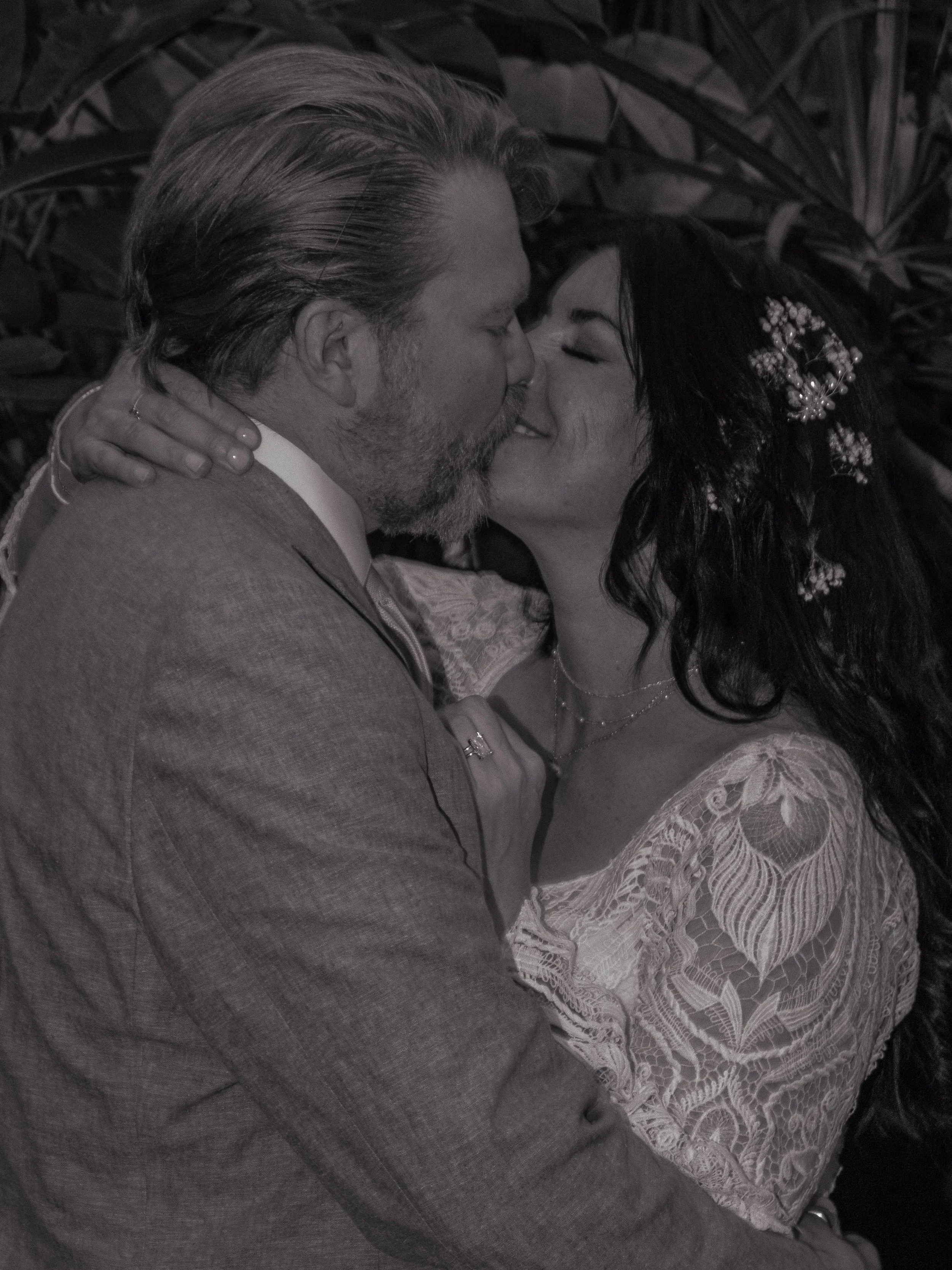 A Black and white photo of a man and a woman sharing a kiss. The man has long, slicked-back hair and a beard, dressed in a suit. The woman has long dark hair with decorative hair accessories, and is wearing lace clothing. Their eyes are closed as they kiss.