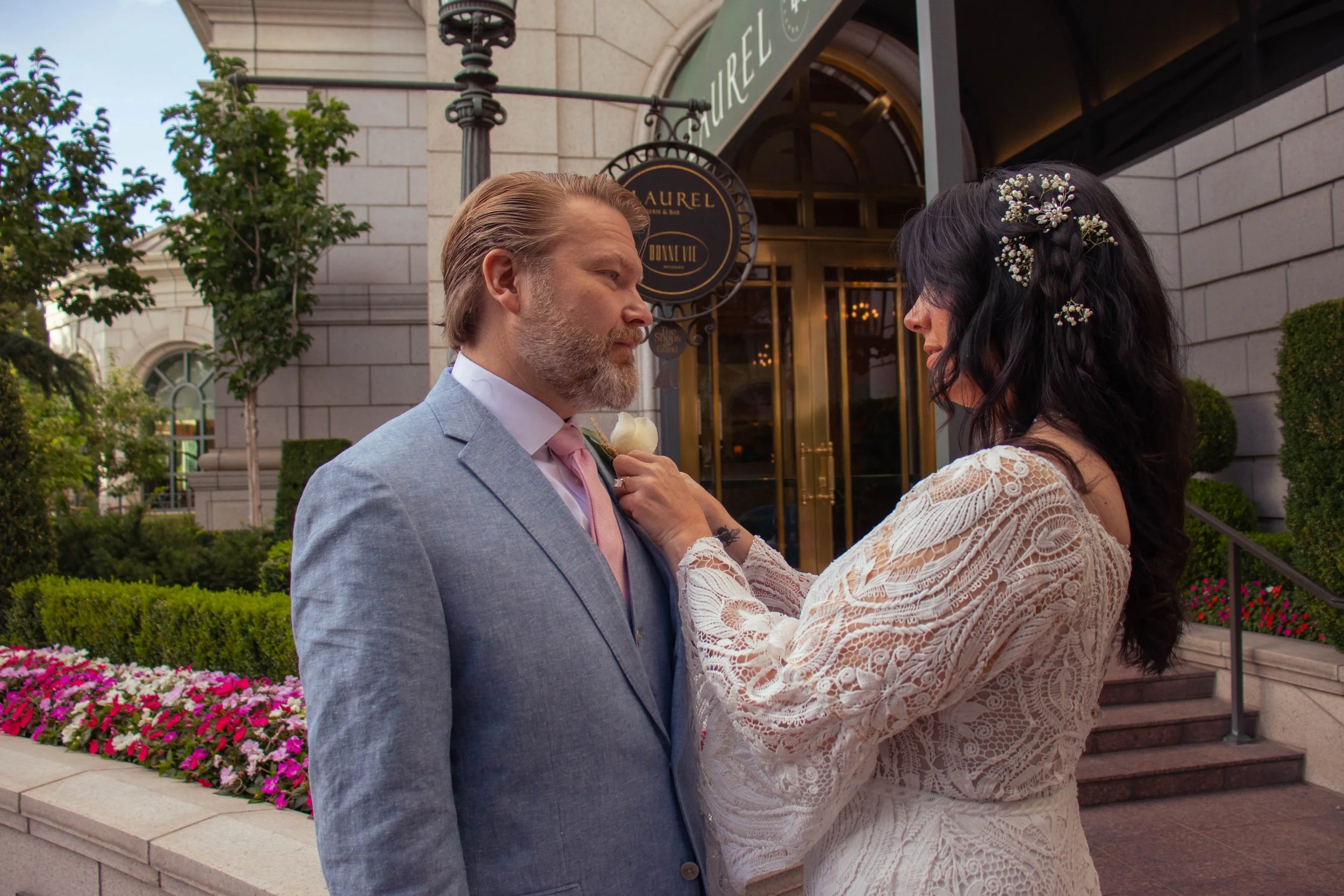 A man and woman facing each other outdoors near a building with signs reading 'Aurel' and 'Bonne Vee.' The man has light-colored hair and a beard, wearing a light blue suit with a pink tie, holding a white rose. The woman has dark wavy hair with smal