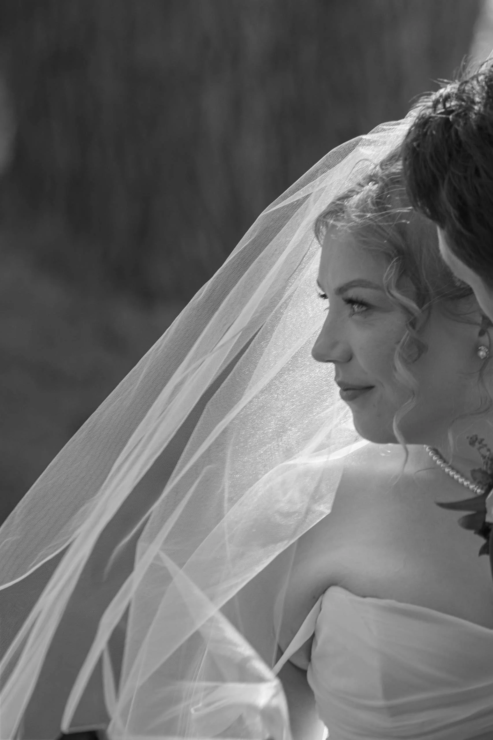 A black-and-white photo of a bride in profile with a long veil, smiling softly, and looking at a man, possibly the groom, who is partially visible.