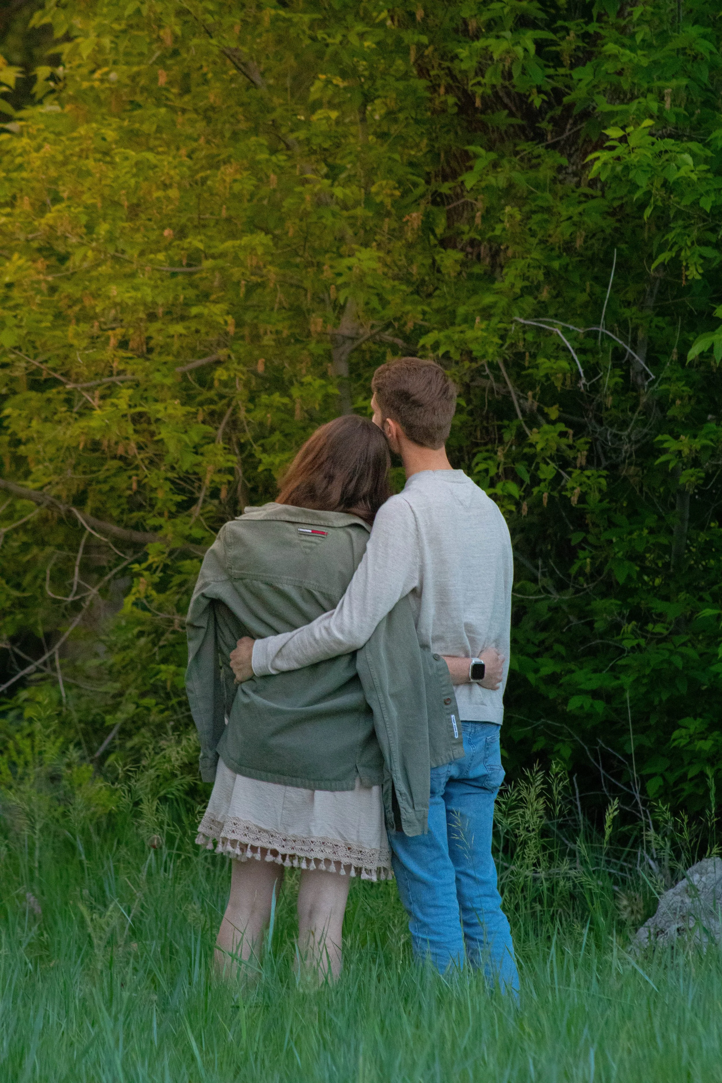A couple stands closely together outdoors in a green, wooded area during daytime, with their backs to the camera. The woman has long brown hair and is wearing a green jacket with a beige skirt underneath. The man has short brown hair and is wearing a