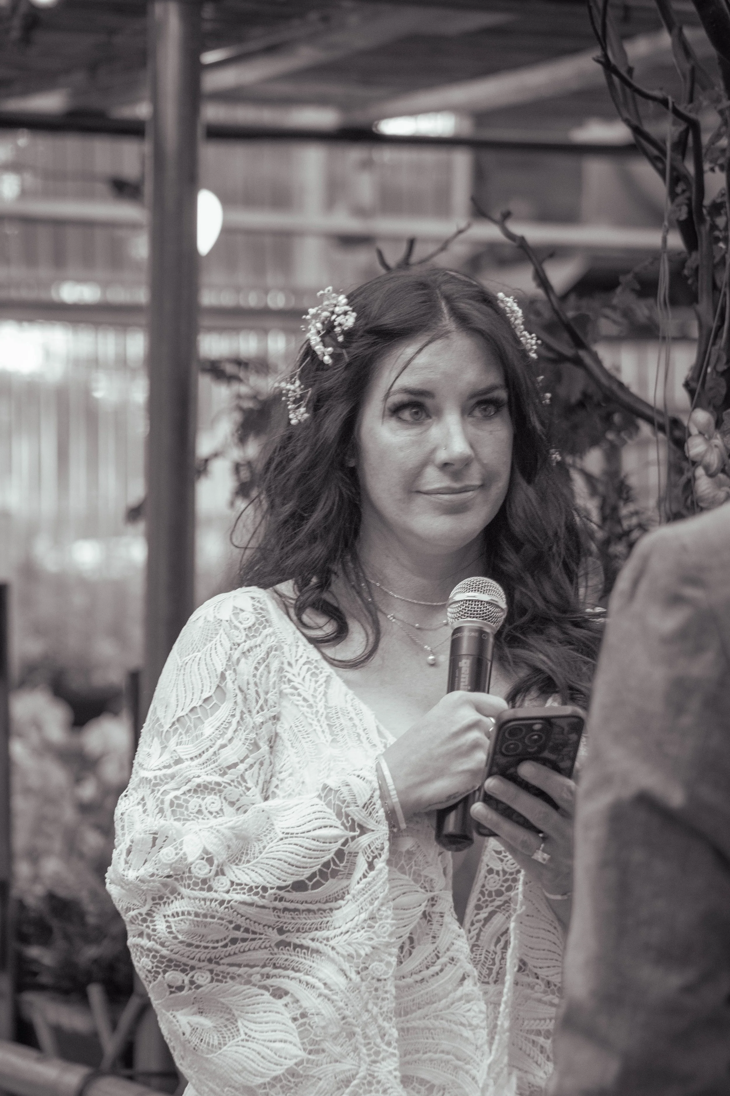 A woman with long dark hair, adorned with small white flowers, is holding a microphone and a smartphone. She is dressed in a lacy white dress and is engaged in a conversation with another person, visible only by their arm and shirt. The background ap