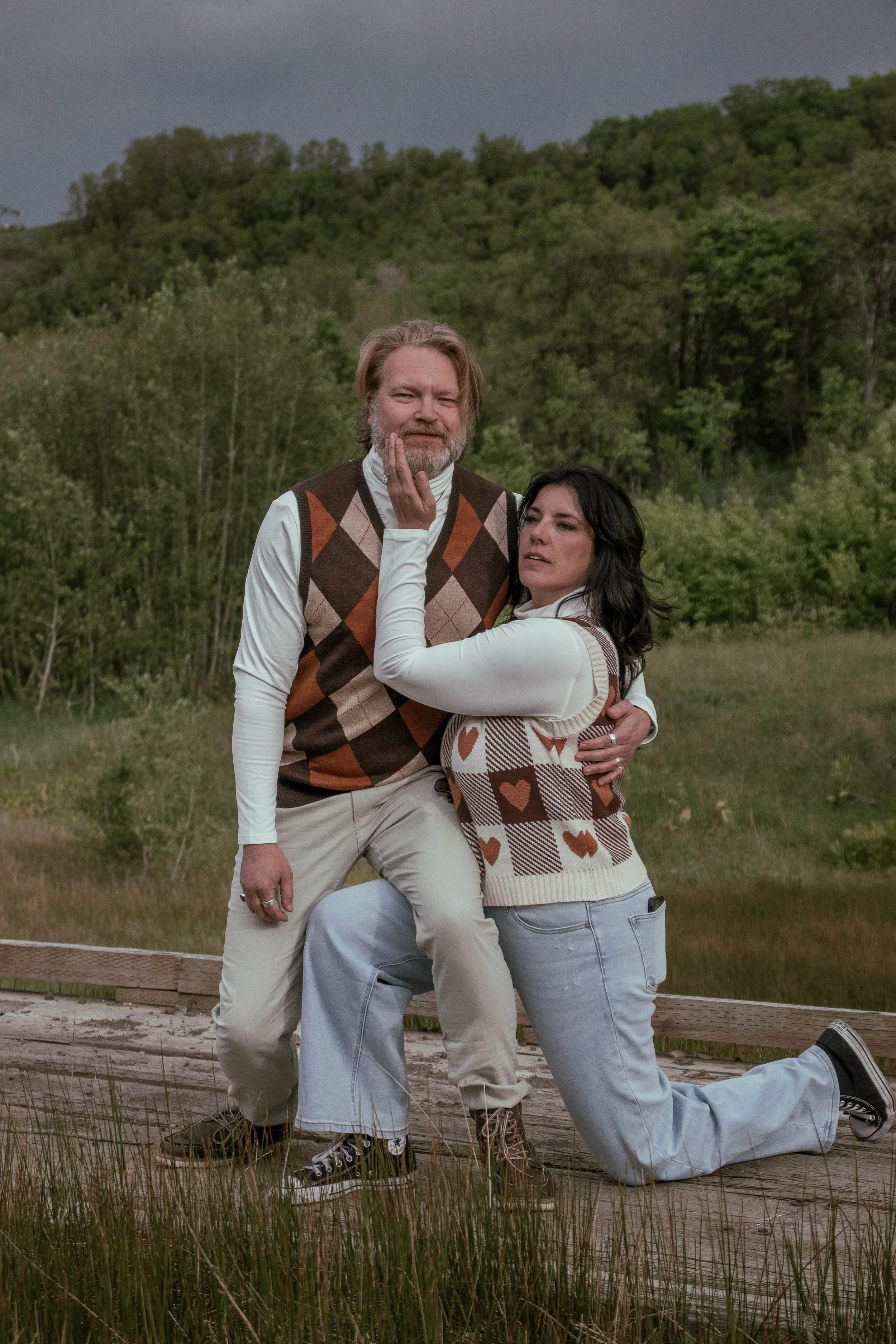A man and woman in outdoor clothing kneel on grass surrounded by tall grass and trees, with a cloudy sky above.