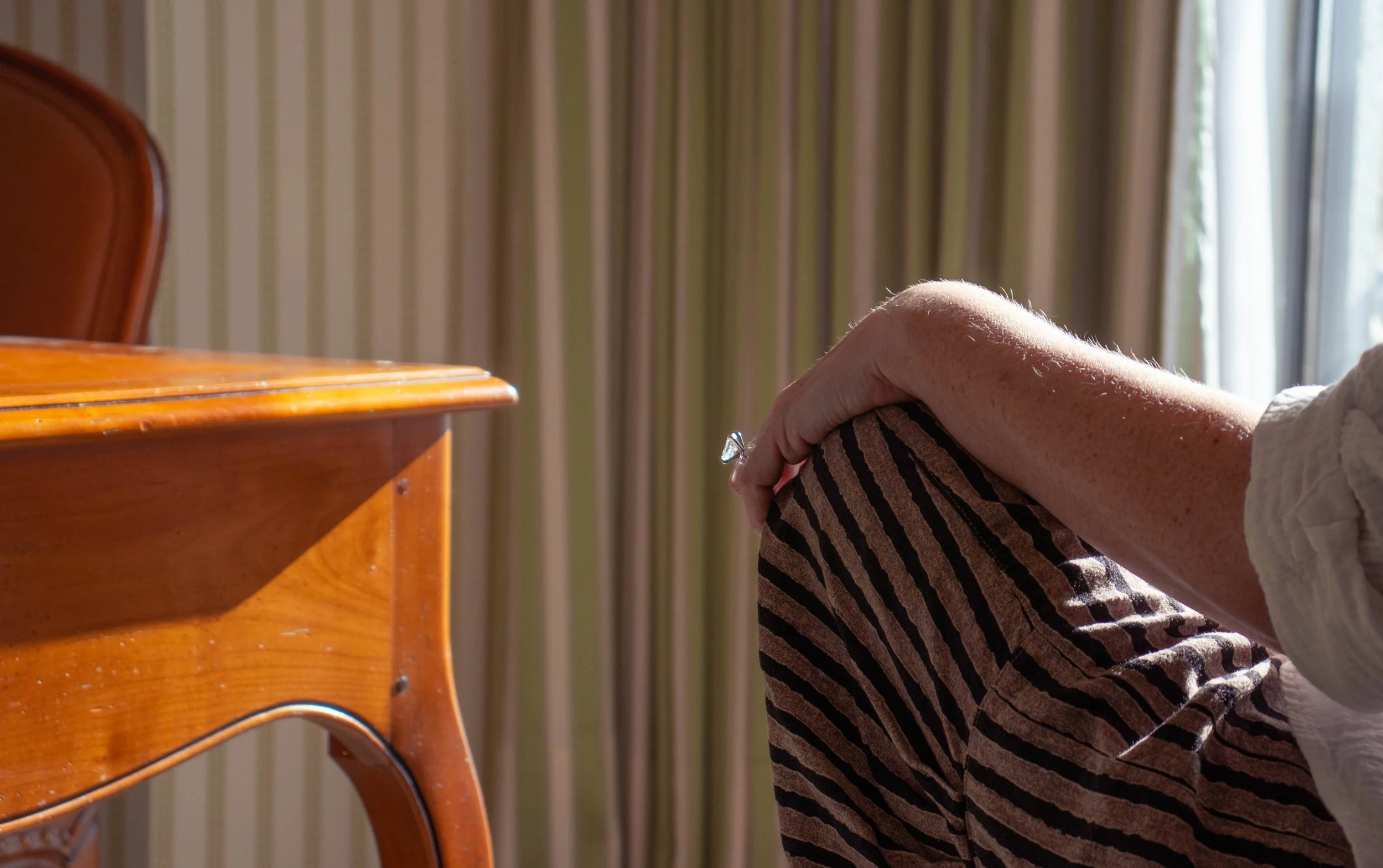 A person sitting on the floor, leaning against a wall next to a window, with a wooden table nearby. The person's face is not visible, and they are wearing a striped skirt or pants.