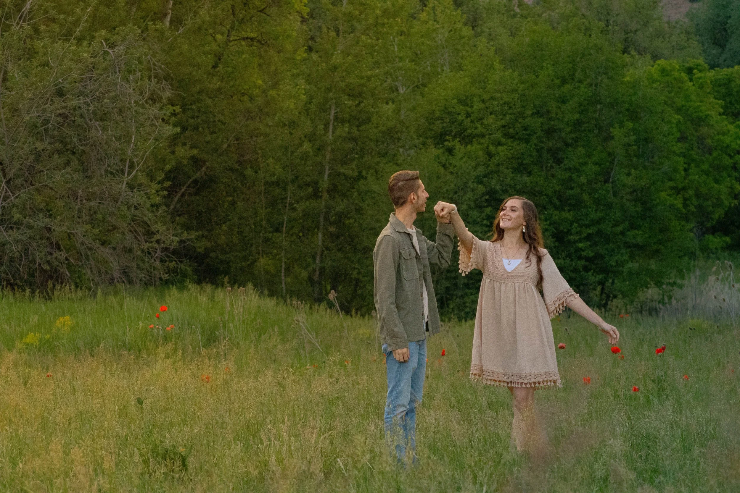 A man and woman standing in a green field with red flowers, holding hands and smiling at each other, surrounded by trees.