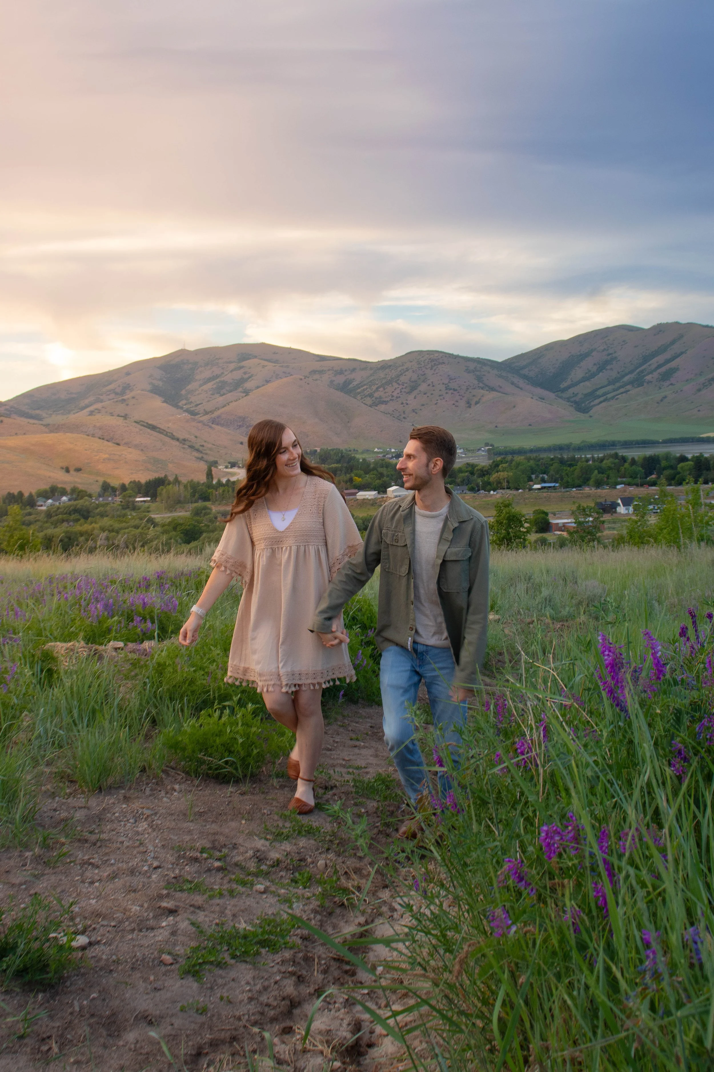 A young couple walking hand-in-hand through a field of purple flowers with mountains in the background during sunset or sunrise, smiling and looking at each other.