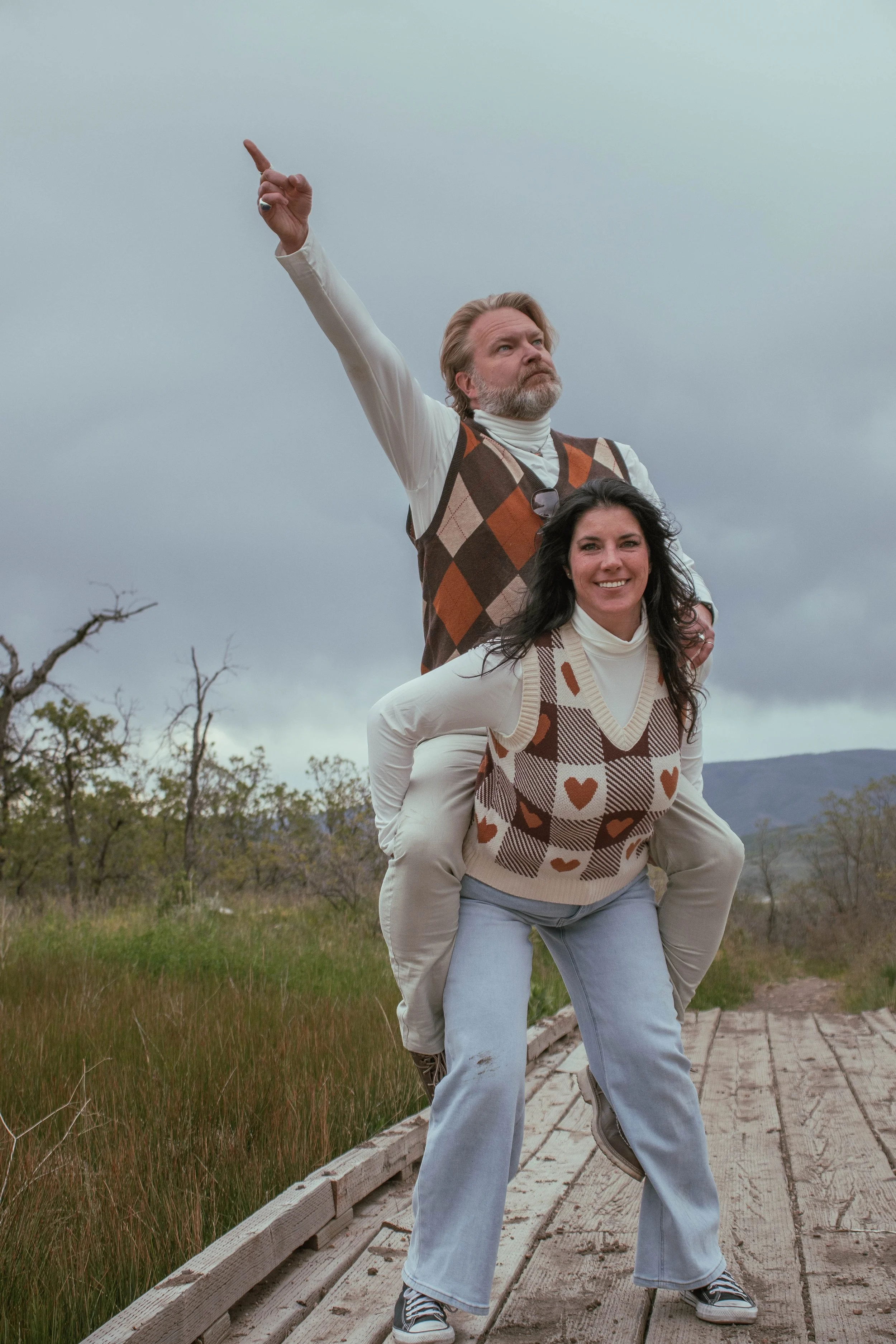A man and woman outdoors on a cloudy day, with the man giving a piggyback ride and the woman smiling. The man is pointing up, and both are dressed in retro-style clothing with argyle patterns. They are standing on a wooden pathway with grass and tree