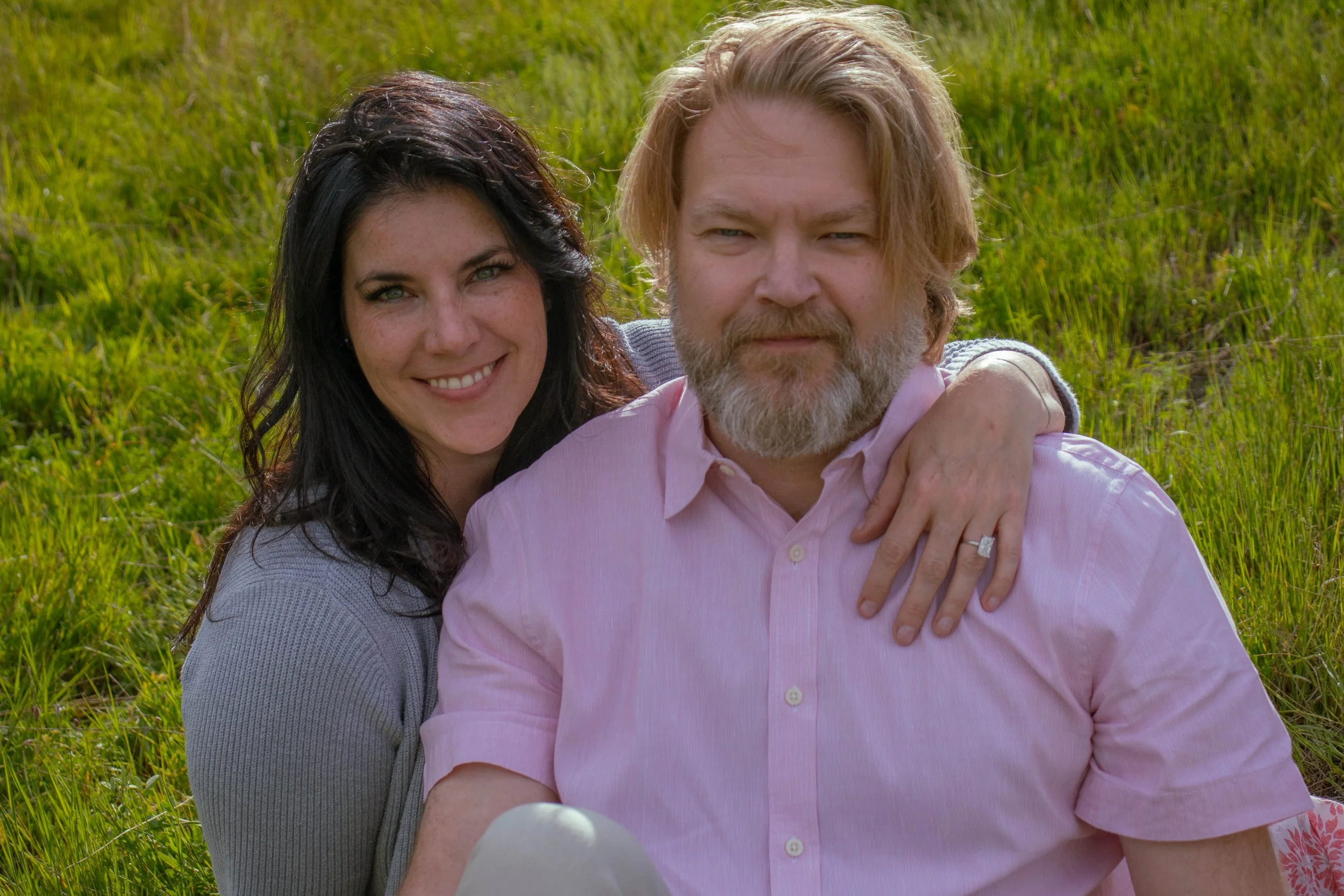 A couple, a woman with long dark hair and a man with long blond hair and beard, sitting close together on grass in a field. The woman is smiling and resting her arm on the man's shoulder, who looks serious. They are outdoors.