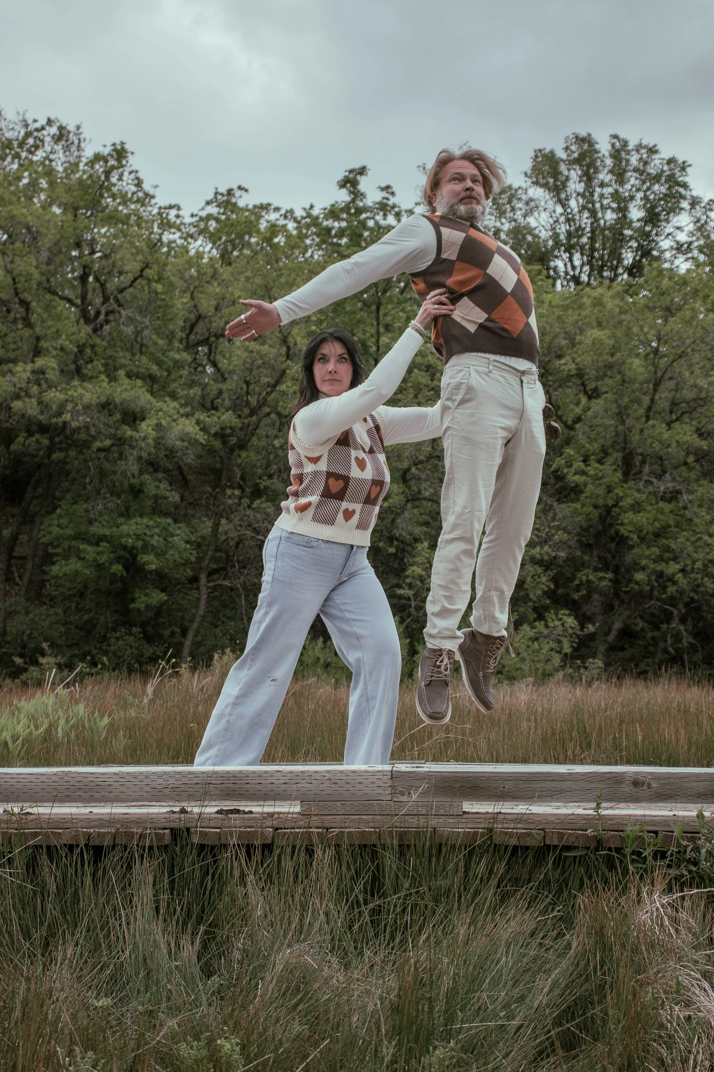 A man with a beard and a woman with dark hair are outdoors on a cloudy day surrounded by trees. The man is dressed in light-colored pants and a vest with an argyle pattern. He is mid-jump with arms extended outward and looking into the distance. The 