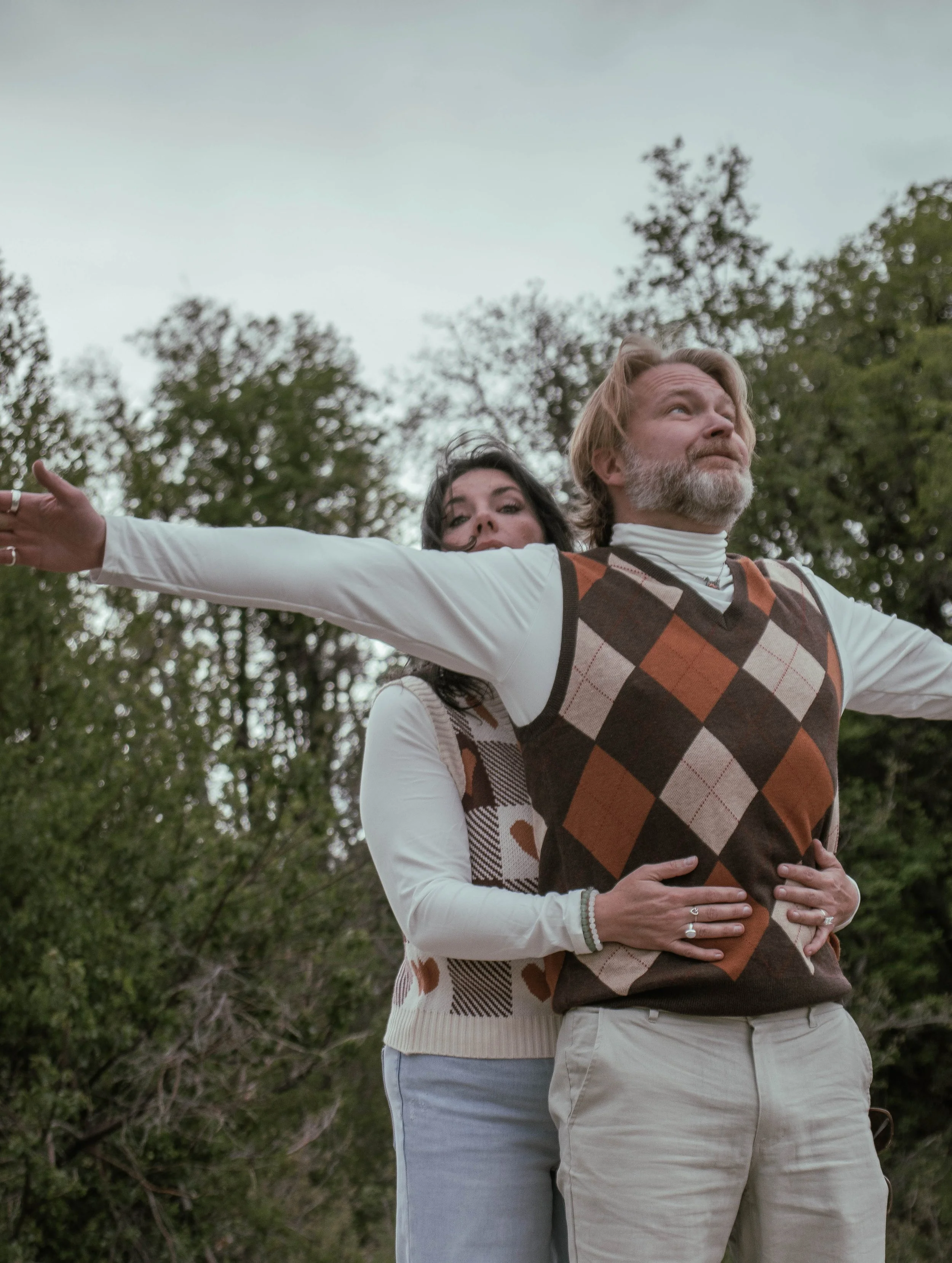 A woman standing behind a man with her arms around his waist in an outdoor setting with trees, while he extends his arms outward wearing a sweater vest with an argyle pattern.