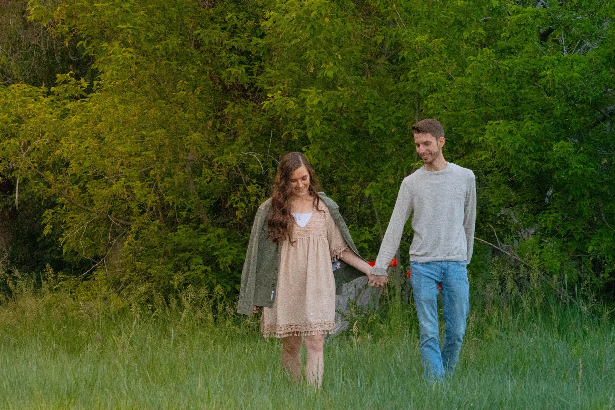 A young man and woman holding hands and walking through a grassy field with green trees in the background, smiling and enjoying each other's company.