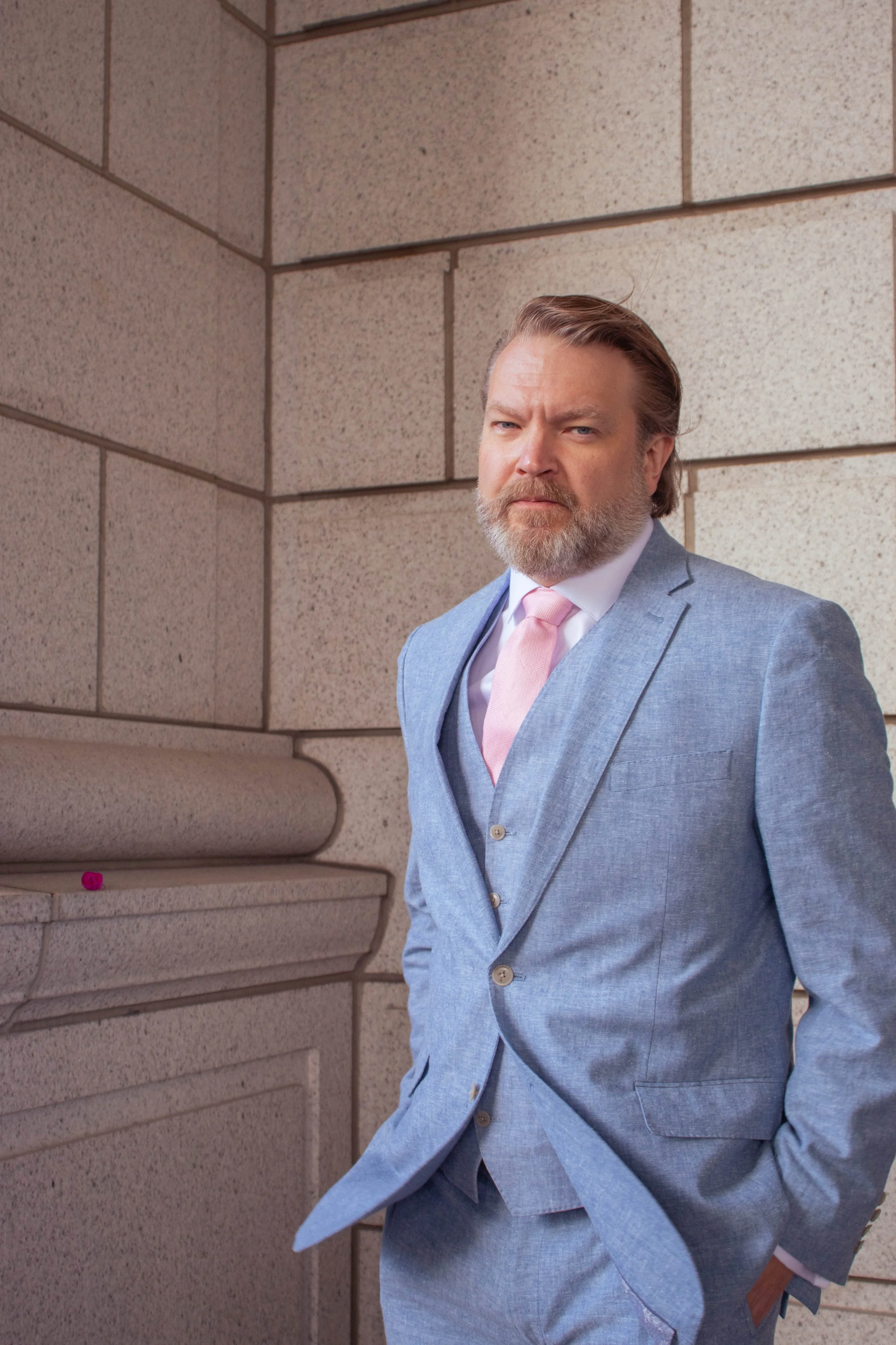A middle-aged man with a beard and styled hair in a light gray suit with a white shirt and pink tie, standing against a brick wall, looking directly at the camera with a serious expression.