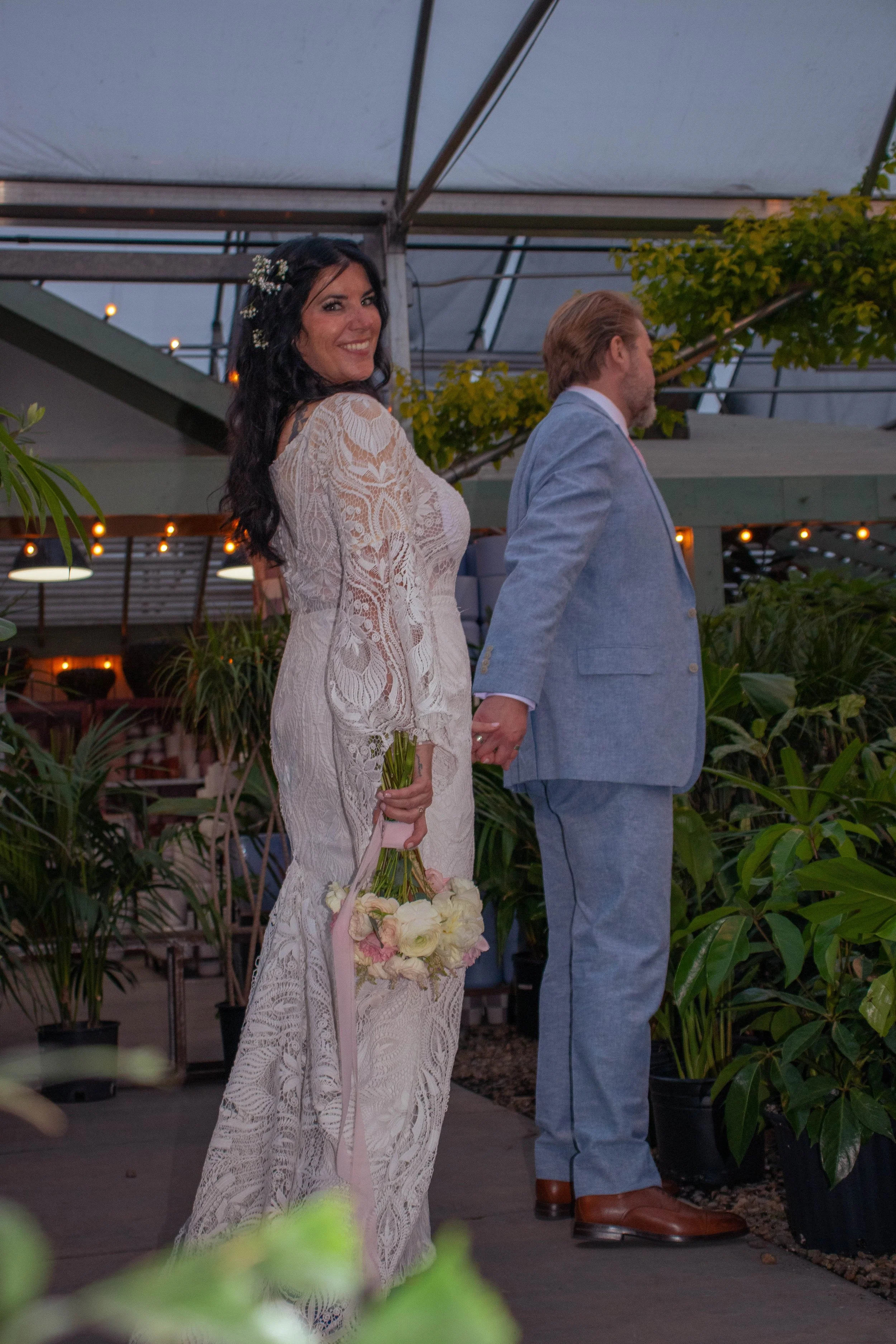 A bride in a white lace dress with floral embellishments and a bouquet of white and pink flowers stands on the left, holding hands with a groom in a light blue suit, during an outdoor wedding ceremony in the evening. Both are facing away from the cam