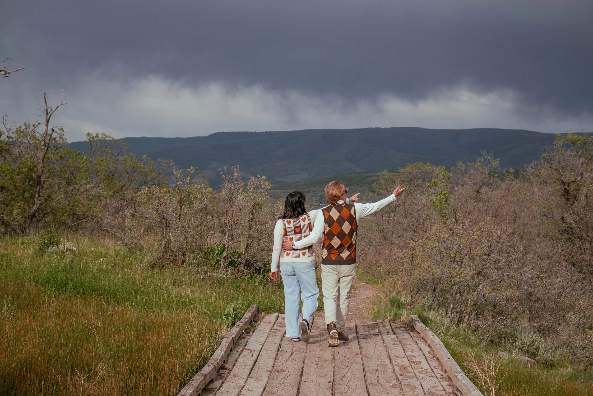 Two people walk hand-in-hand on a wooden bridge through a nature landscape with trees and distant mountains, under dark stormy clouds.