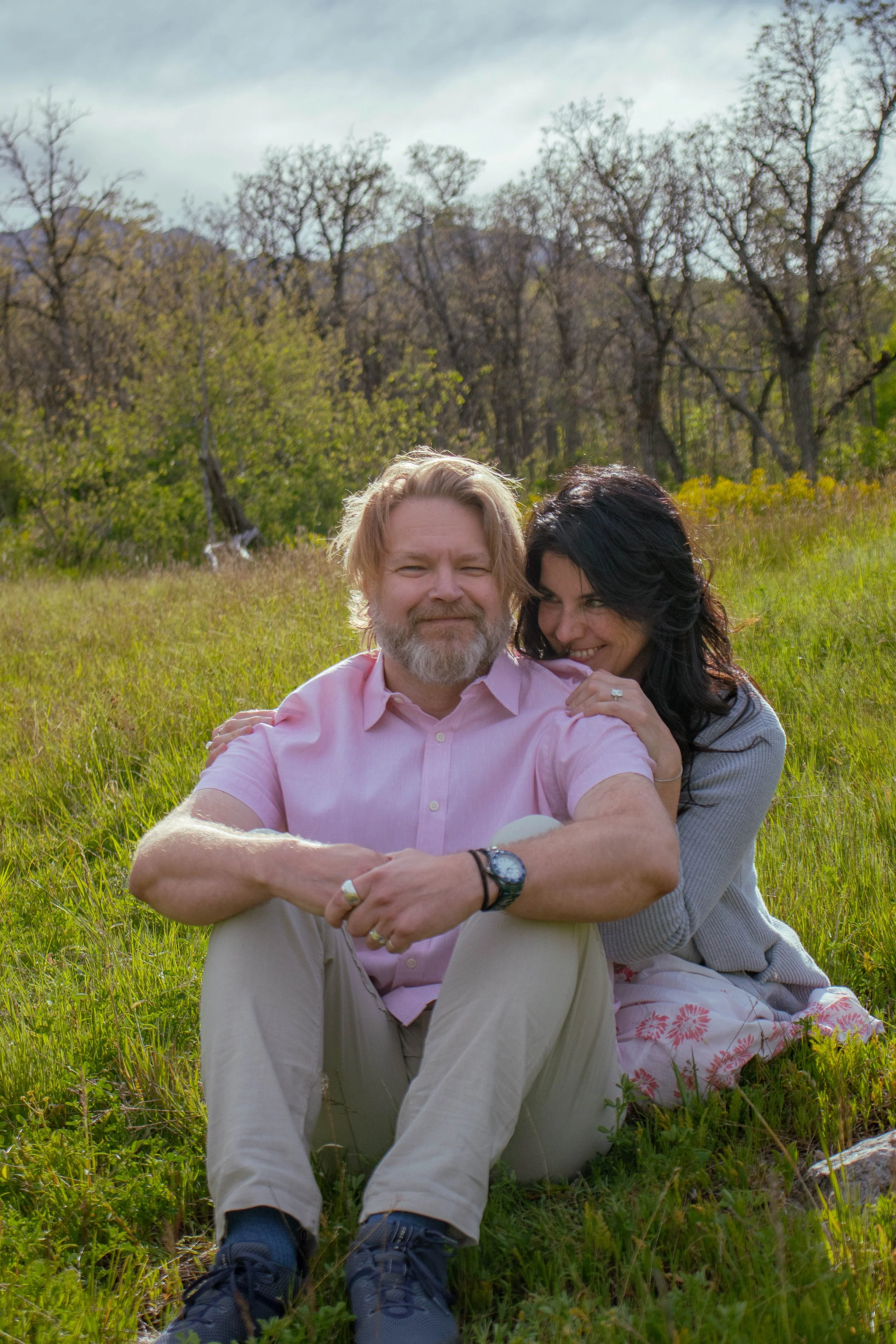 A man with a beard and glasses sitting on the grass, wearing a pink shirt, with a woman with dark hair smiling and embracing him from behind, in a field with trees in the background on a cloudy day.