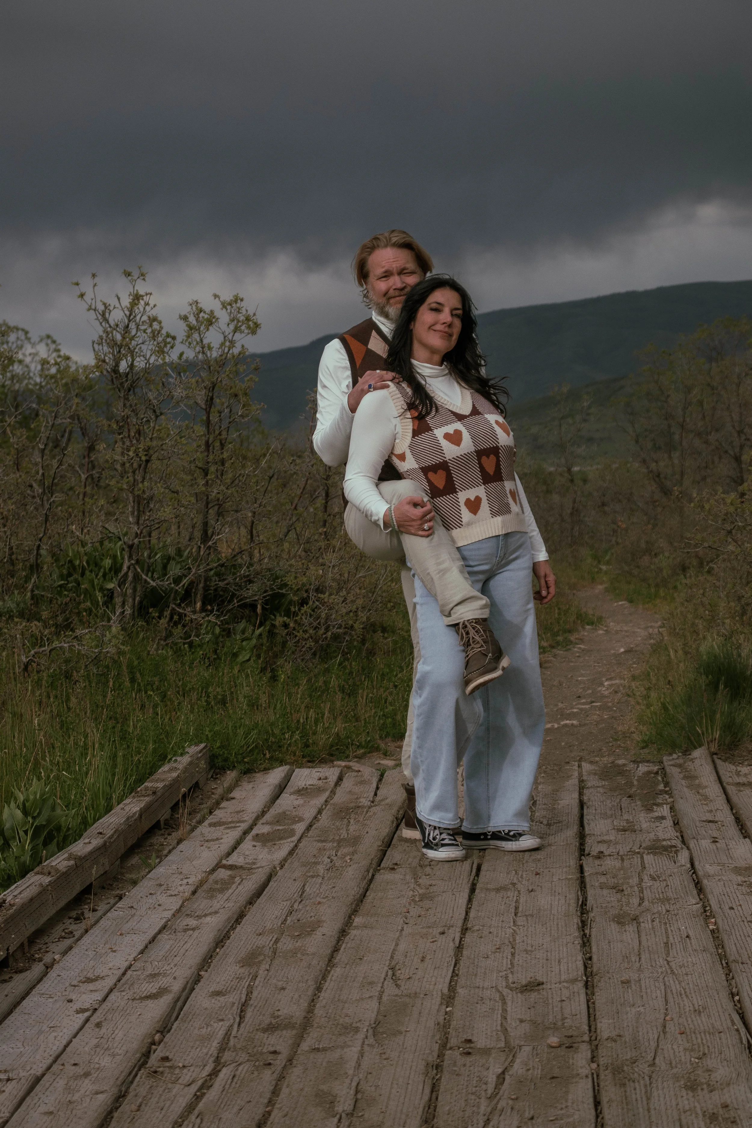 A couple standing on a wooden bridge outdoors with a stormy mountain landscape in the background. The man is carrying the woman on his back, and they are both smiling.