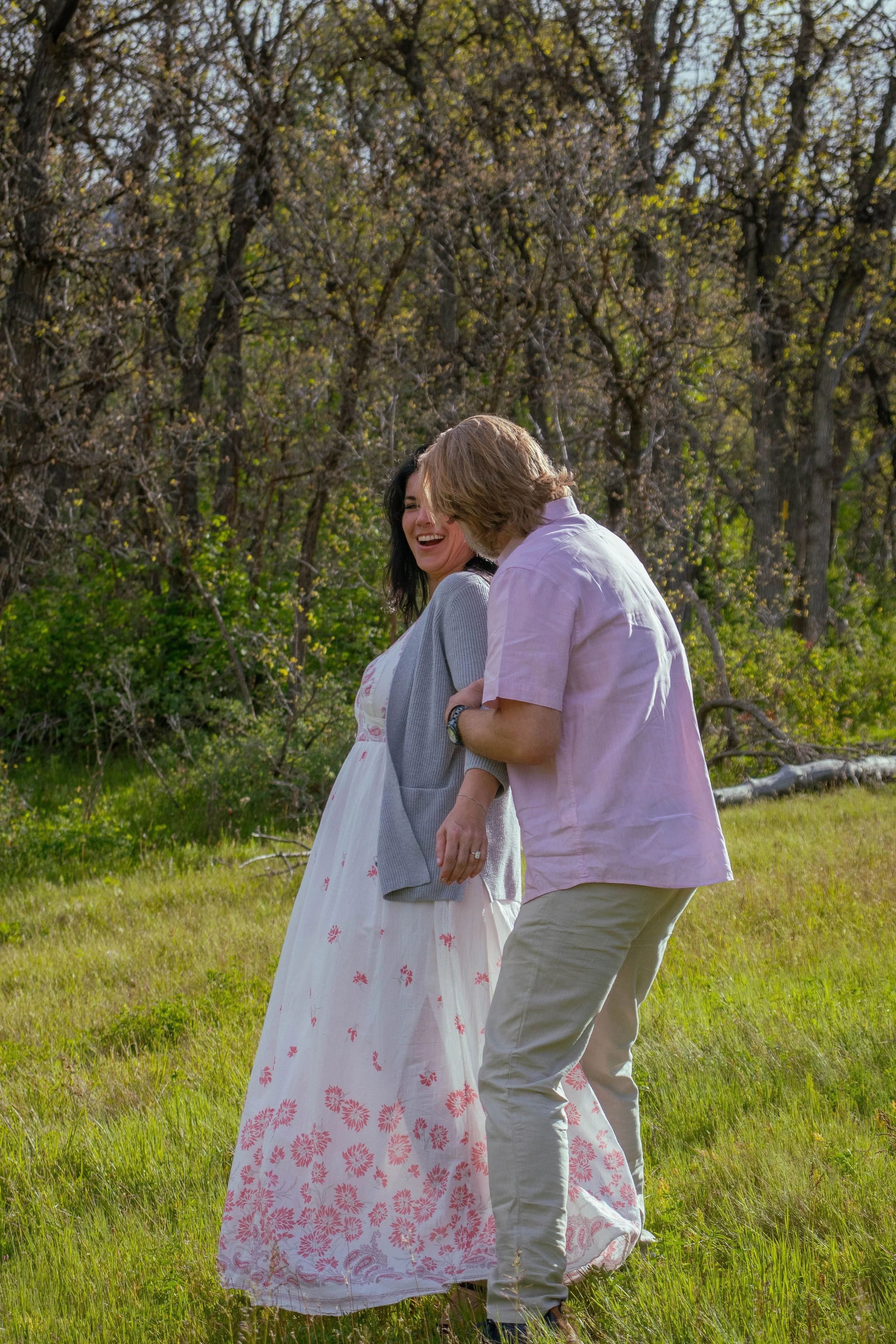 A couple standing on grass in a park, smiling and holding hands, with trees in the background on a sunny day.