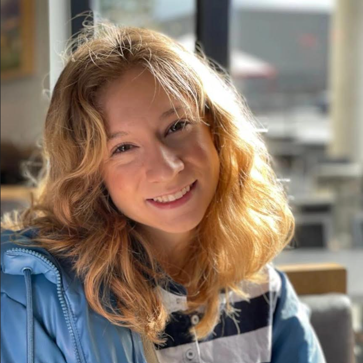 A young woman with wavy red hair smiling at the camera, sitting indoors near a window with sunlight streaming in.