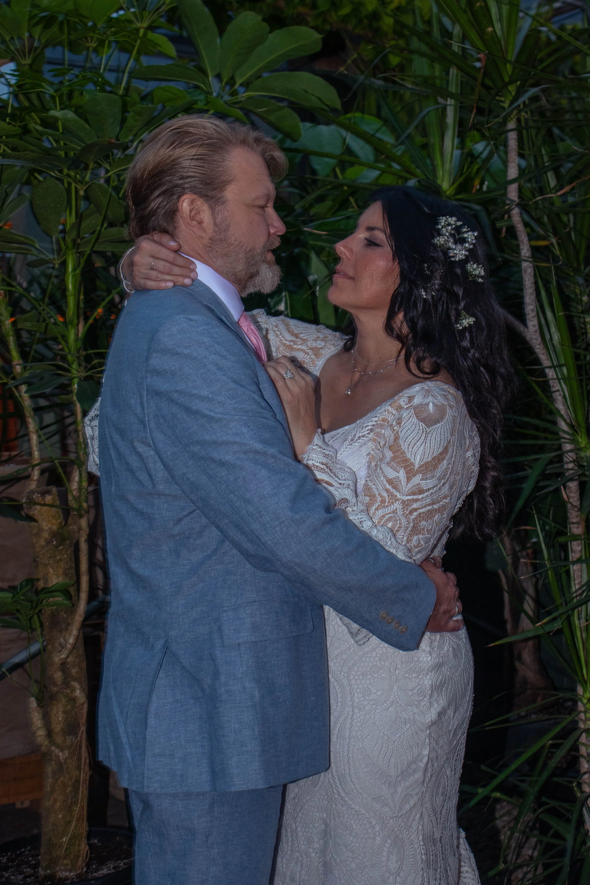 A man and woman are dancing closely outdoors among leafy plants or trees at dusk, with the man in a gray suit and the woman in a white lace dress with flowers in her hair.