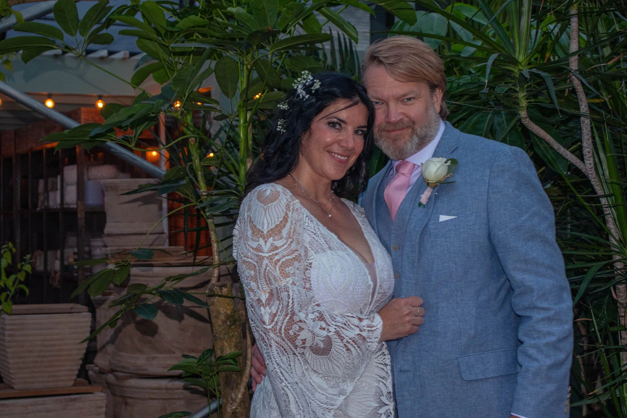 A couple dressed in wedding attire standing close together outdoors surrounded by green plants and trees, smiling at the camera during the evening.