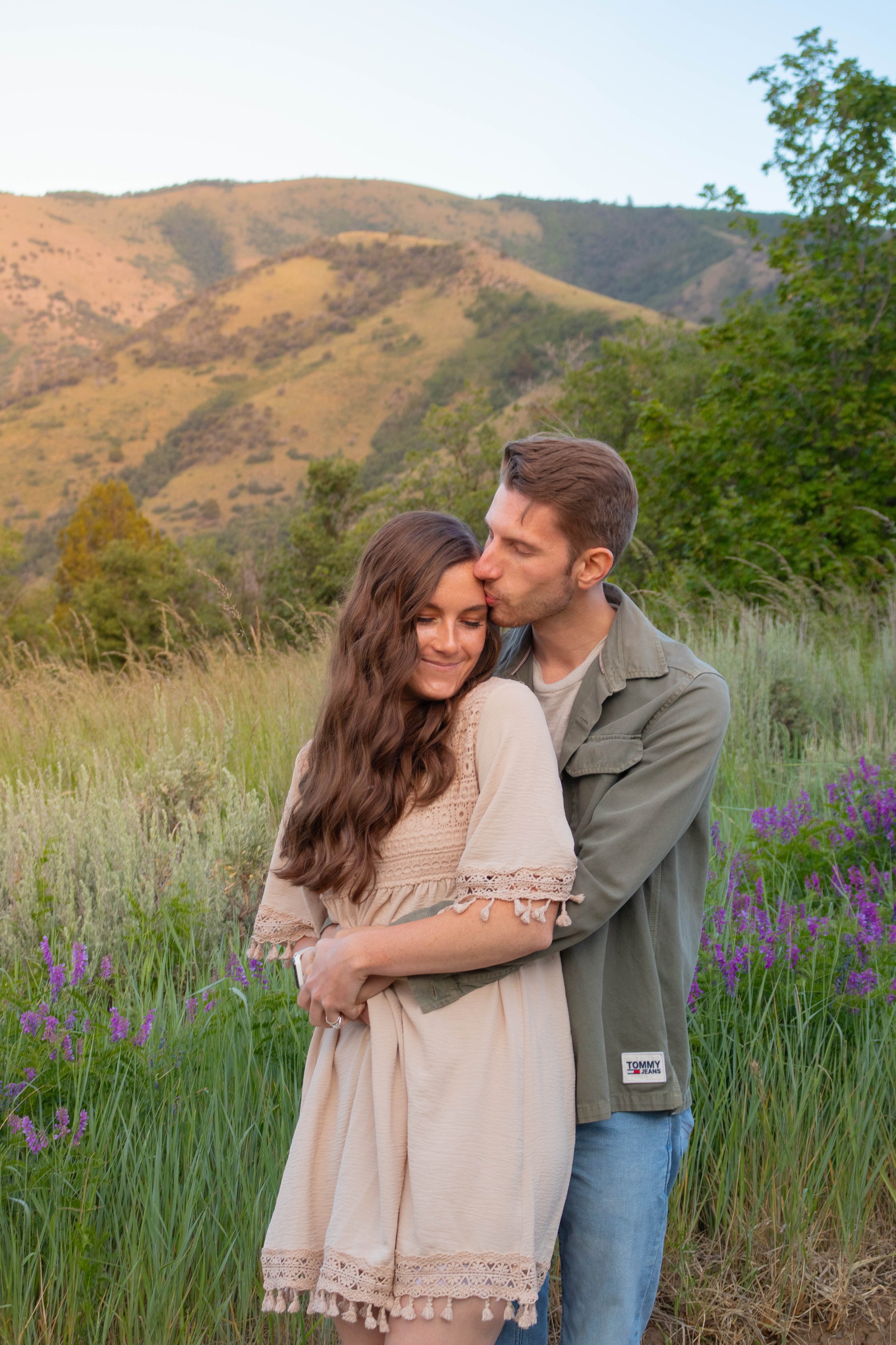 A young couple embracing in a field of tall grass and purple flowers, with green trees and mountains in the background, during late afternoon or early evening.