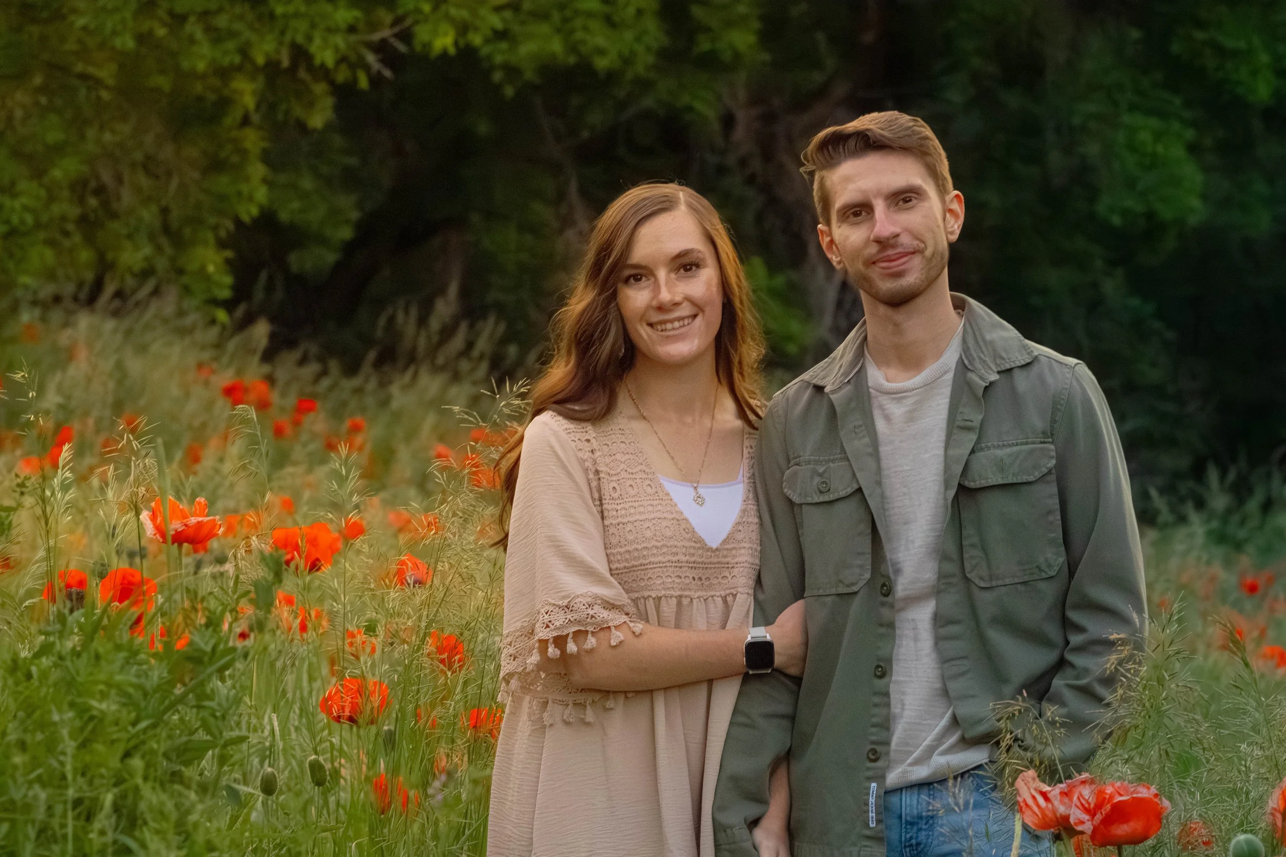 A smiling wife and man standing in a field of red poppies with green trees in the background.
