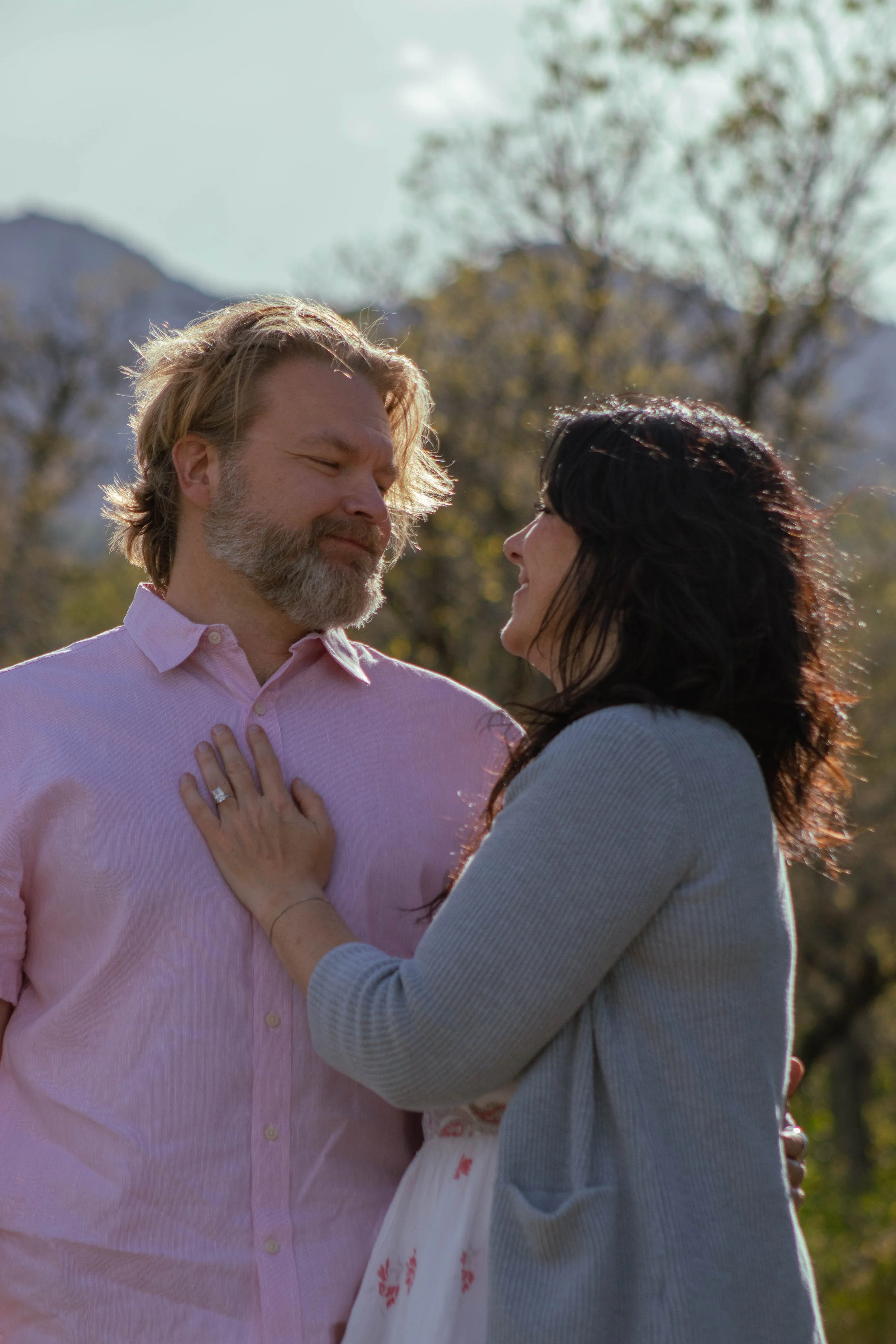A man with long blond hair and a beard wearing a pink shirt, and a woman with dark hair wearing a gray sweater, stand close together outdoors with mountains and trees in the background. The woman is touching the man's chest and they are smiling at ea