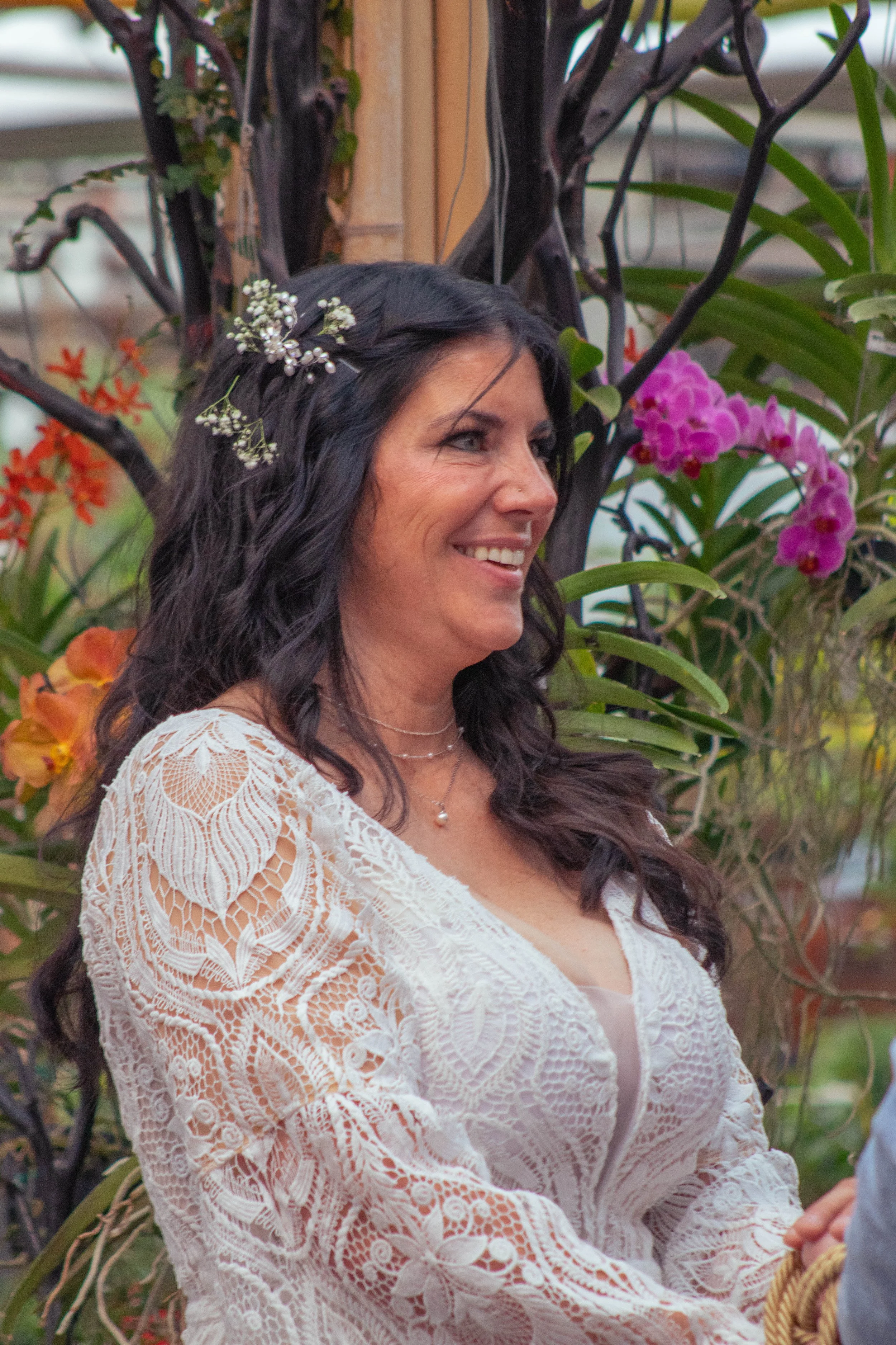 A woman with long dark hair styled with small white flowers, smiling while sitting outdoors among colorful flowers and green plants, wearing a white lace dress and a pearl necklace.