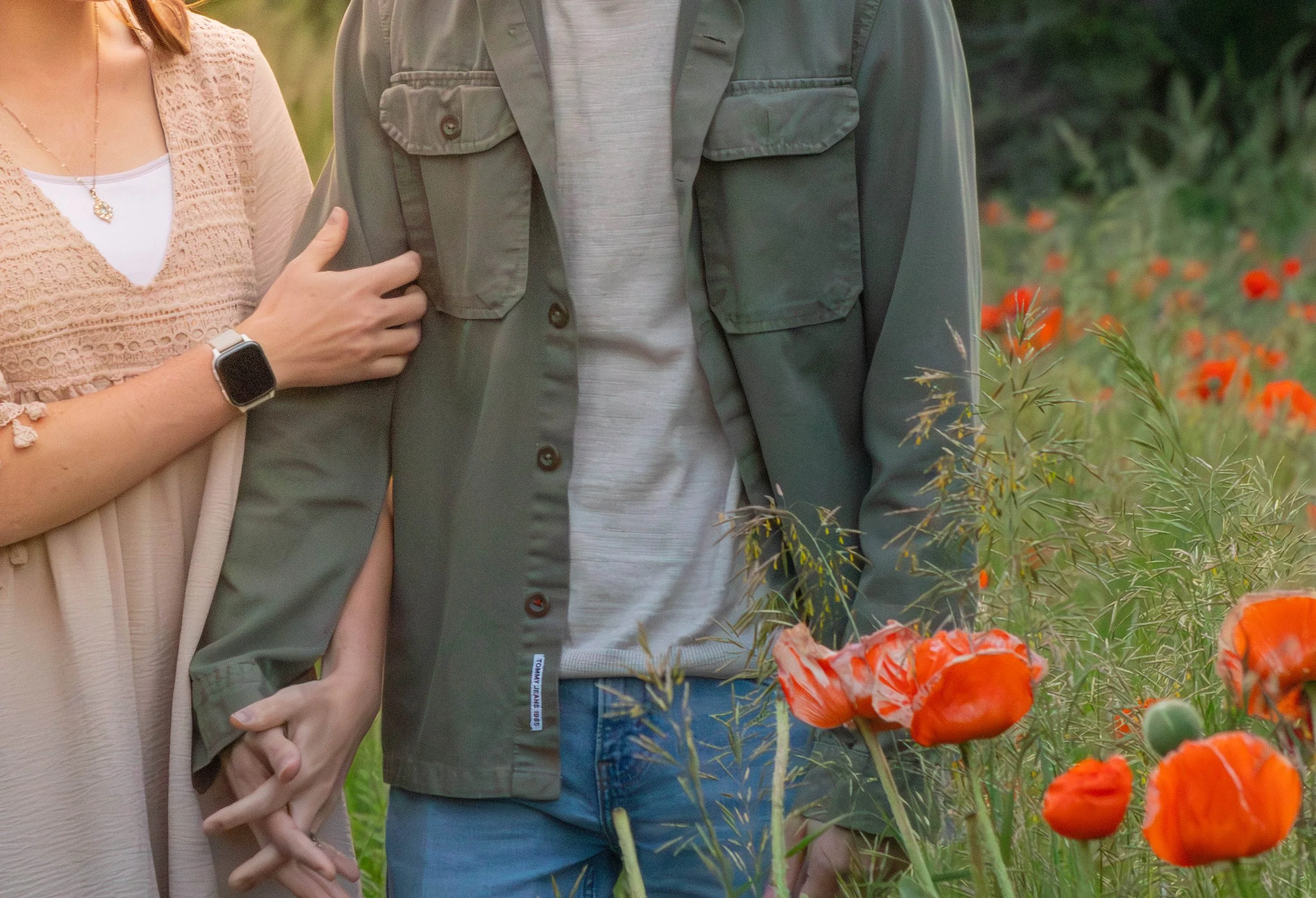 Close-up of a couple holding hands, standing in a field of orange poppies, with the woman's arm around the man's shoulder, in an outdoor setting with greenery.