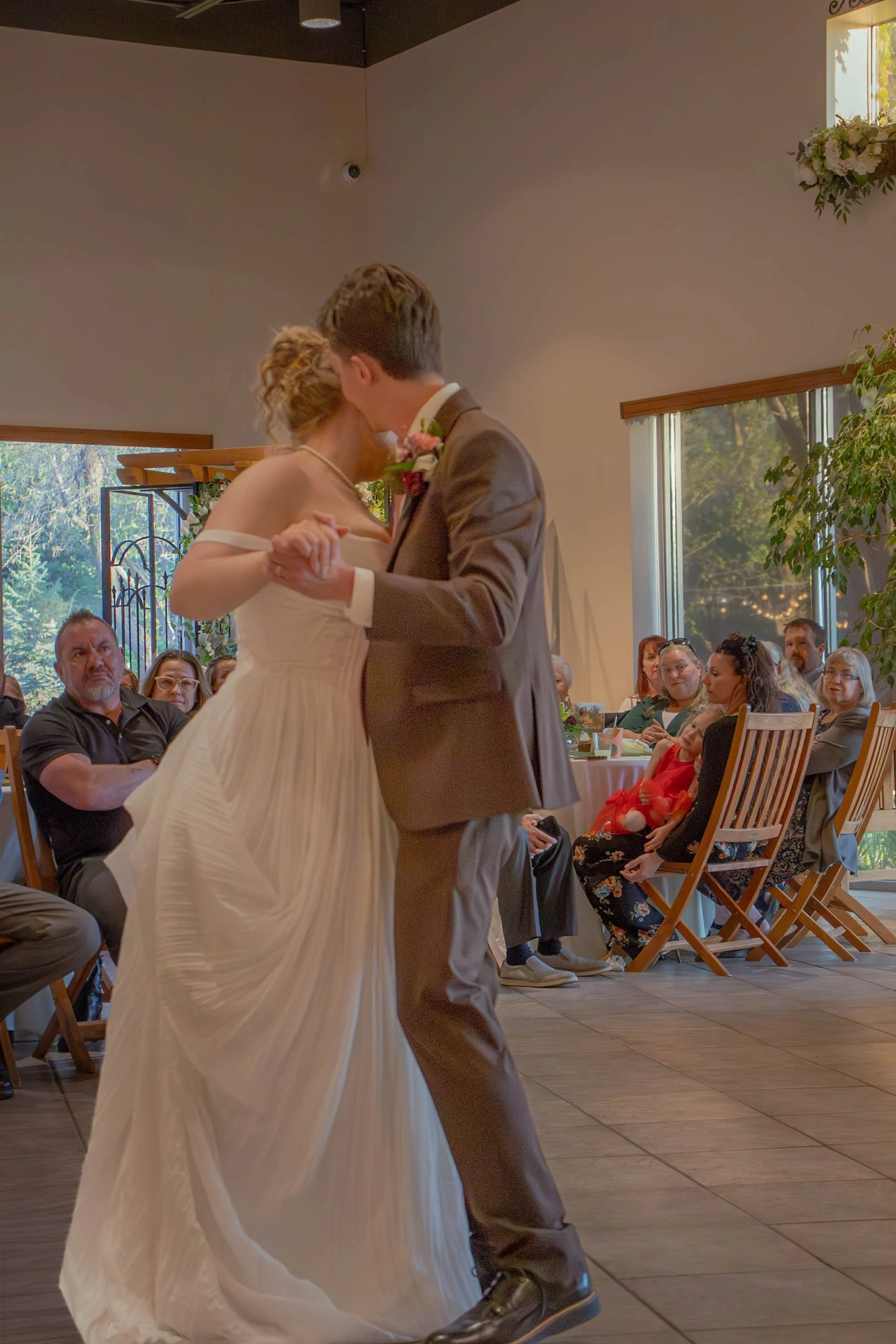 A bride and groom sharing a dance at their wedding reception, surrounded by seated guests in a warmly decorated indoor venue with large windows and greenery outside.